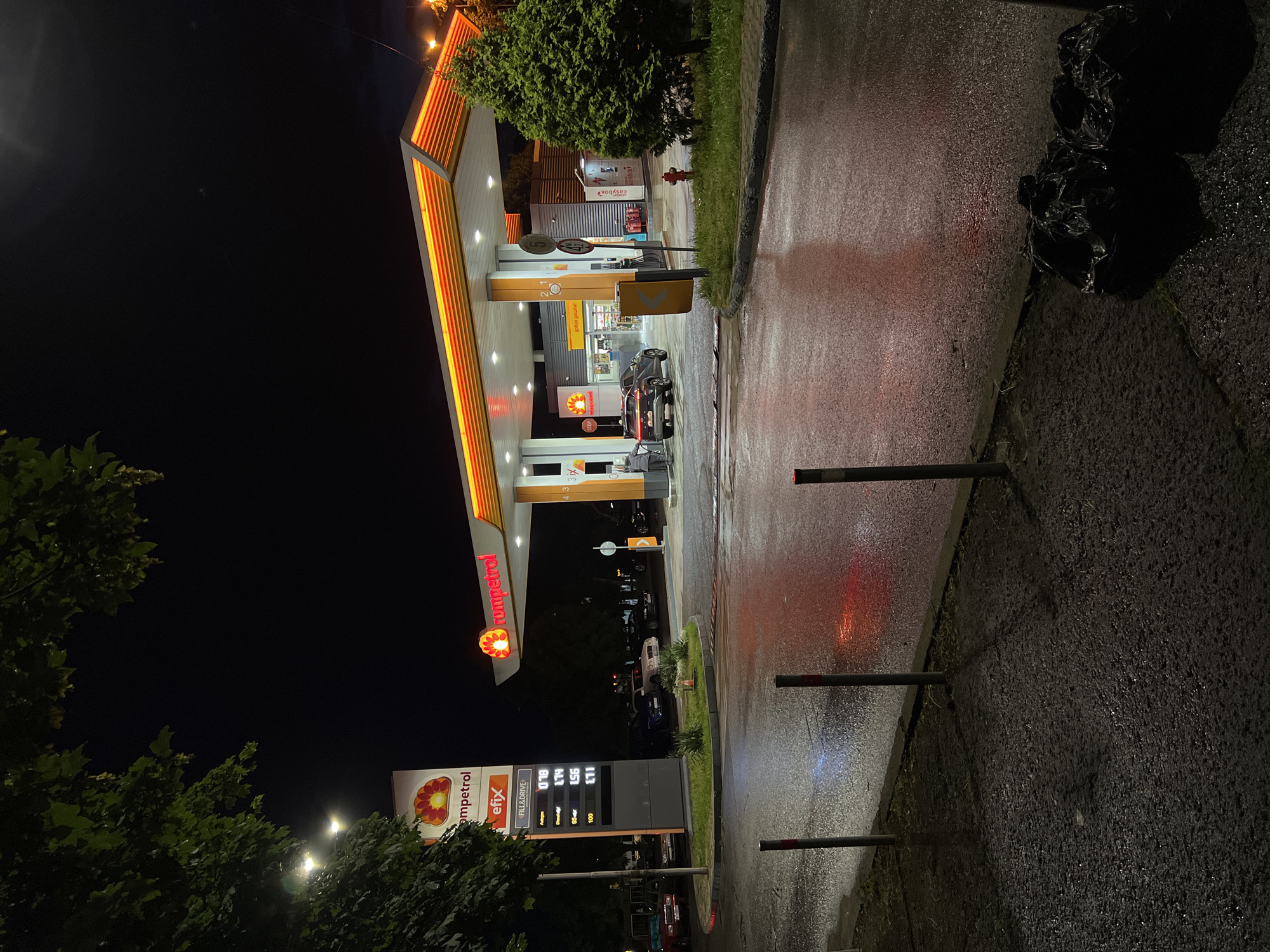 The image captures a nighttime scene at a Rompetrol gas station in Burgas, Bulgaria. The station is well-lit, with prominent Rompetrol signage and fuel price displays showing prices for Fill&Drive, 95, 95+, and 100 fuel types. Vehicles are parked at the gas pumps. The adjacent sidewalk and road are wet, likely from recent rain, and reflect the station's lights. Black garbage bags sit beside the sidewalk. Across the street is a sign with “easybox” printed on it, and several parked cars. Trees are seen on the far left of the image. The sky is black. The orientation of the image is rotated 90 degrees.