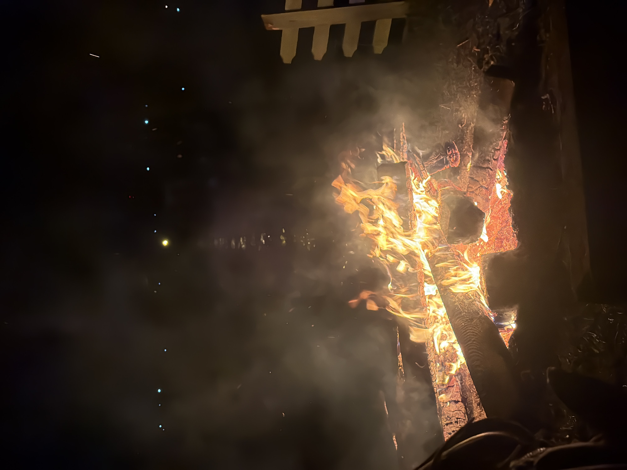 The image depicts a bonfire at night, possibly in a rural or semi-rural setting, as indicated by the presence of lights in the distance that could be from a town or settlement. There is a white, horizontal wooden fence visible on the right side of the frame. The bonfire itself is substantial, with large logs burning brightly, emitting significant heat and smoke. The flames are vibrant orange and yellow, casting a warm glow. In the foreground, there are blurry shapes that suggest people are present, seated or standing around the fire. One of these shapes on the left appears to be an arm or shoulder with dark clothing. The overall atmosphere is one of a nighttime gathering around a fire. The location is identified as Noviy, Russia, though specific landmarks within Noviy are not identifiable from the image alone.