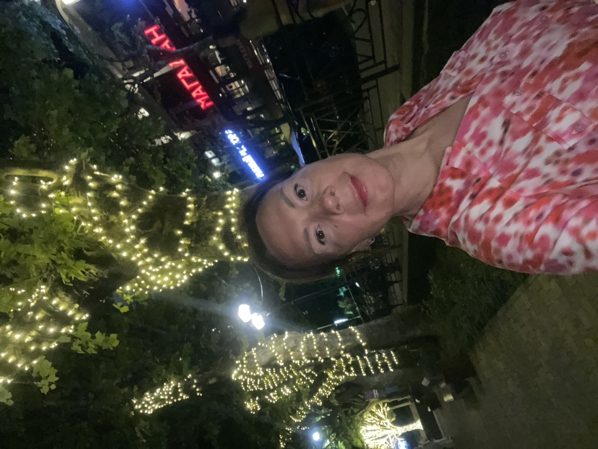 A woman is standing outdoors at night, posing for a selfie. She is wearing a pink and white tie-dye shirt. In the background, trees are decorated with strings of warm white lights. A building with neon signs, one in red that reads "МАГАЗИН" (MAGAZINE), is visible behind the trees. Streetlights are also present, casting light on the scene. The ground appears to be paved, suggesting an urban environment. The overall impression is of a festive or decorated street, possibly in a commercial area. The image was taken in the city of Sochi, Russia.