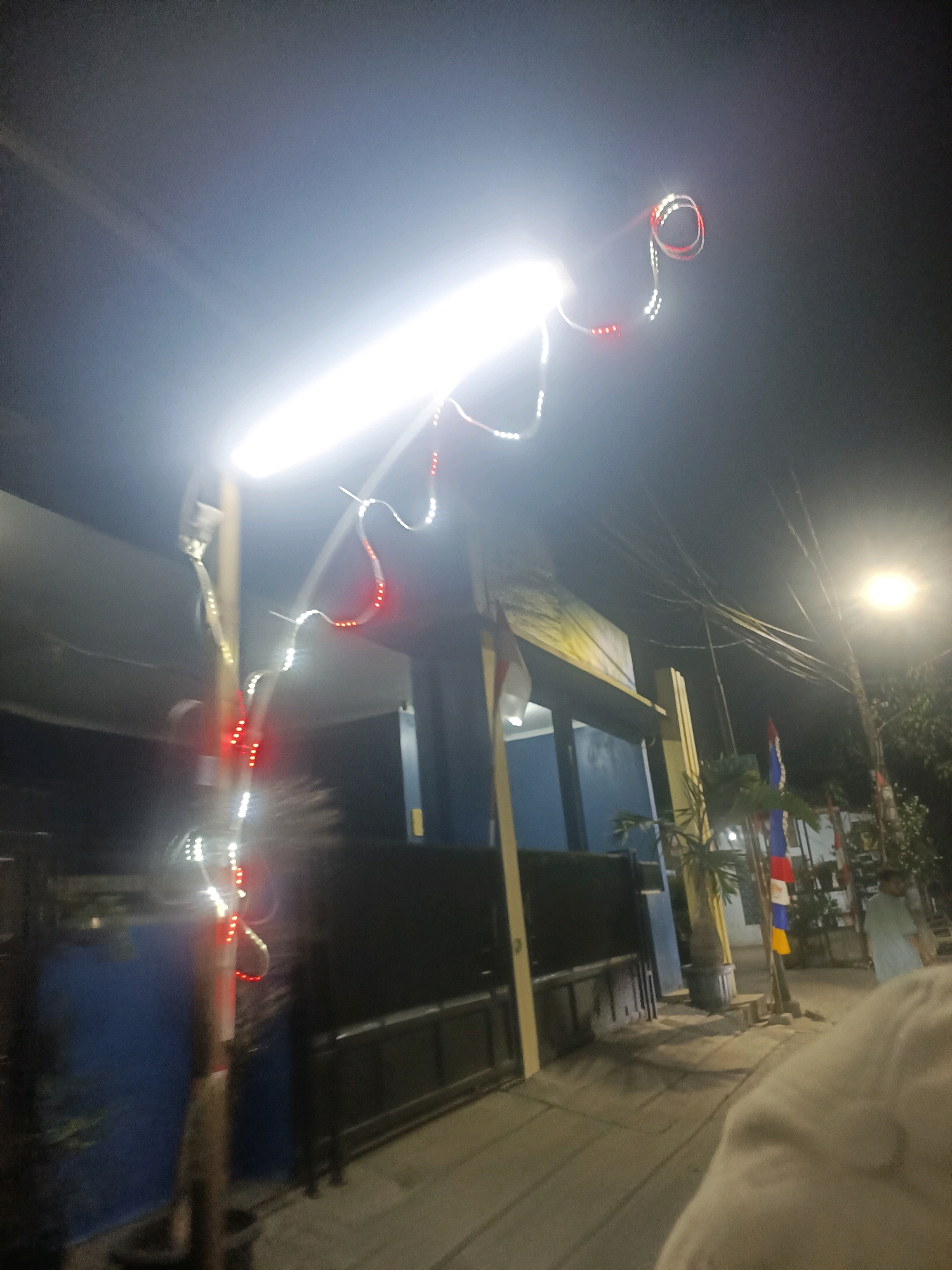 The image depicts a street scene at night in Telagaasih, Indonesia. A bright fluorescent light illuminates the foreground, casting a strong glare. Red and white string lights are draped around a pole and appear to be decorative. In the background, a building with a dark facade and a blue section is visible. Several flags, including one with red and white colors, are planted near the building. Power lines are strung across the sky, and a street lamp emits light to the right of the frame. A person in a light-colored garment is partially visible in the lower right corner, appearing to be moving away from the camera. The ground is paved with concrete, and shadows are cast by the various elements in the scene. The overall impression is that of a decorated street at night, possibly for a celebration or event.