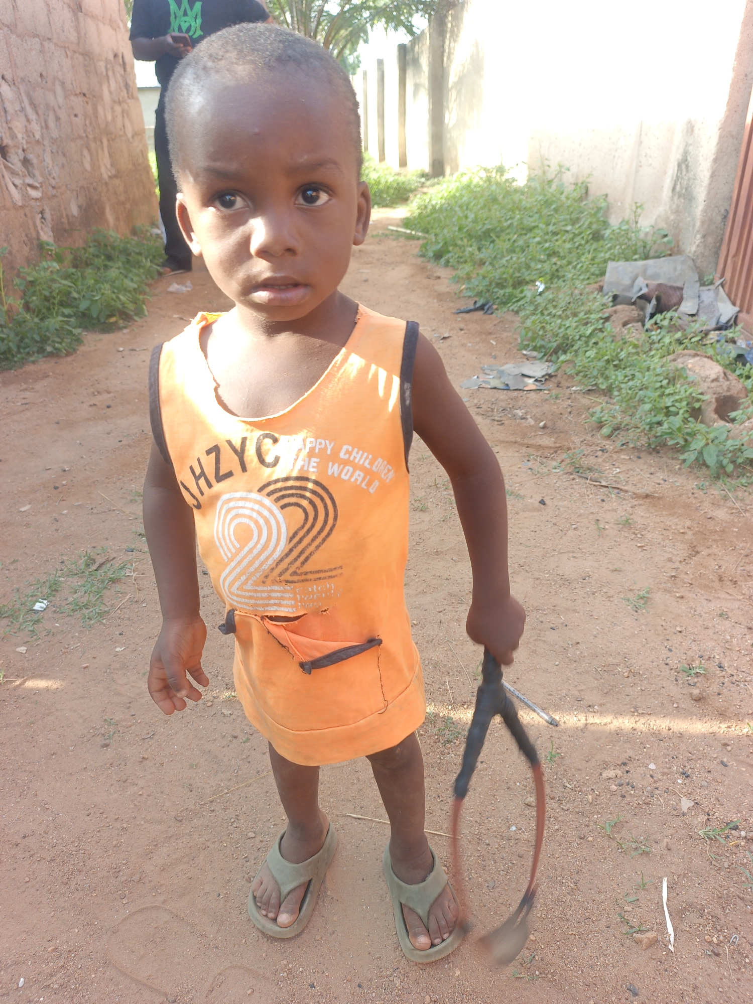 A young child stands on a dirt path in Akwanga, Nigeria. The child is wearing an orange shirt with black trim and a graphic design that says "ZHZYC HAPPY CHILDREN THE WORLD", and green flip-flops. The child holds a makeshift toy consisting of a rubber band stretched around a forked stick. The path is lined by walls and vegetation on either side. A man wearing a black shirt is partially visible in the background on the left.