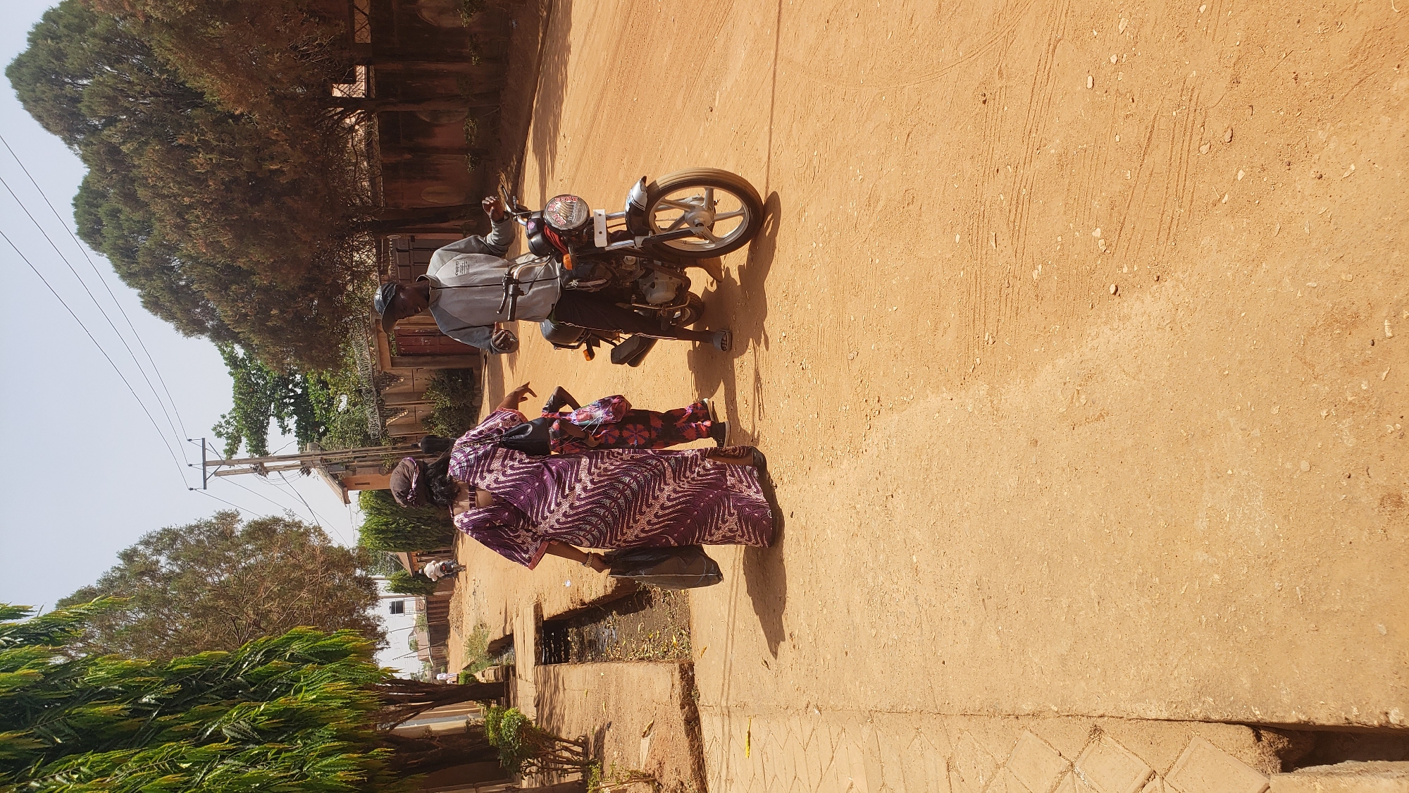 The image shows a dusty street scene in Sabon Tasha, Nigeria. A man wearing a light gray sweatshirt and a dark cap is sitting on a motorcycle. He is facing the viewer's right. To the left of the motorcycle, a woman in a long, patterned purple dress and a child in a patterned red dress stand facing the man. A drain runs along the edge of the road, to the left. Trees line the street, providing partial shade. Buildings are visible in the background.
