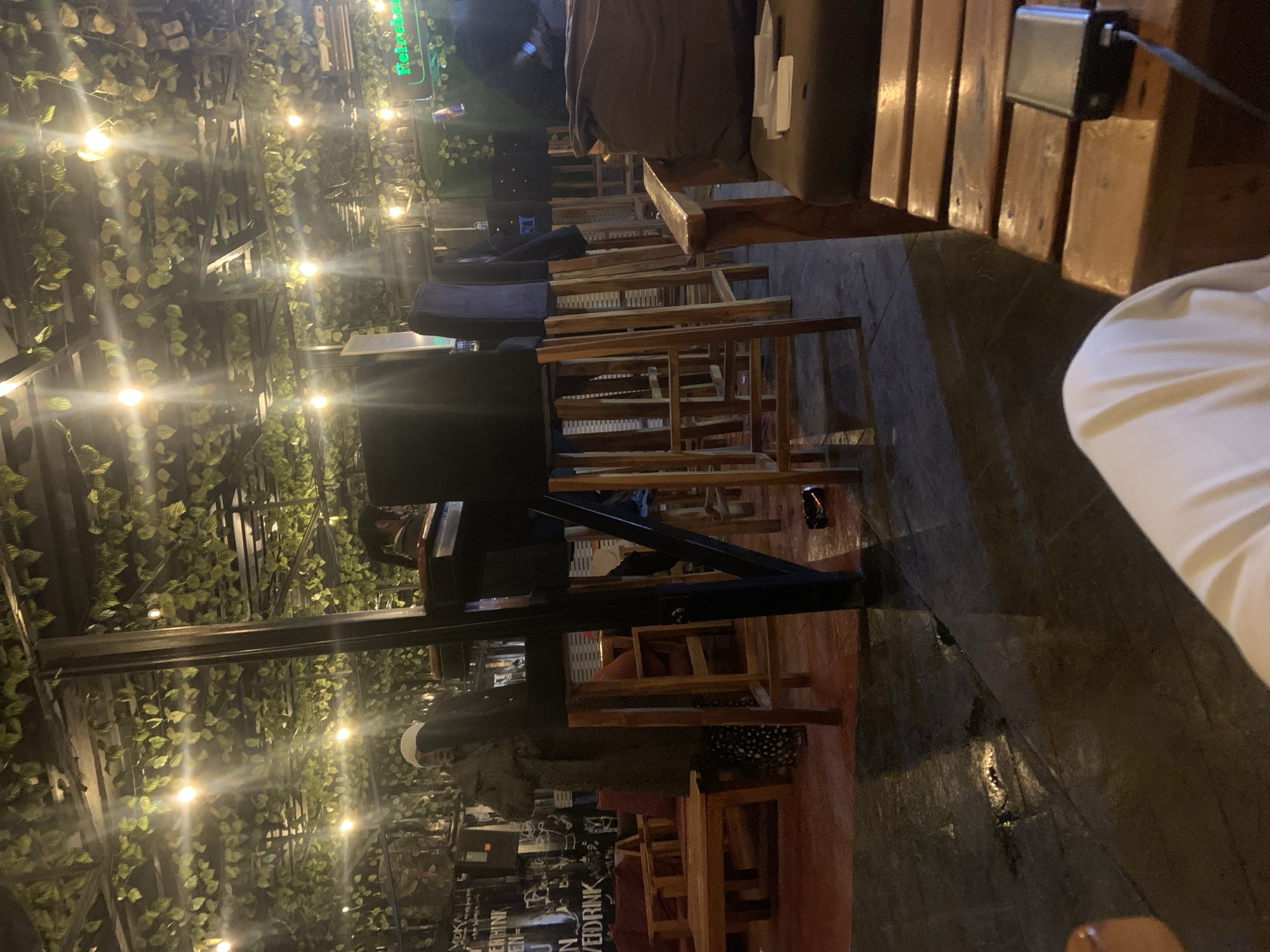 The image depicts an indoor seating area, possibly a bar or restaurant, with a bohemian-style decor. The ceiling is adorned with artificial green foliage and strings of warm-toned lights, creating a cozy ambiance. Several wooden bar stools with dark upholstered seats are arranged around a counter, suggesting a place for drinks and socializing. In the background, a person in a long coat and patterned trousers stands near a wall covered with graffiti and posters. The floor appears to be wet in some areas, reflecting the overhead lights. A Heineken sign is visible in the upper right portion of the frame. The scene evokes a relaxed and casual atmosphere.