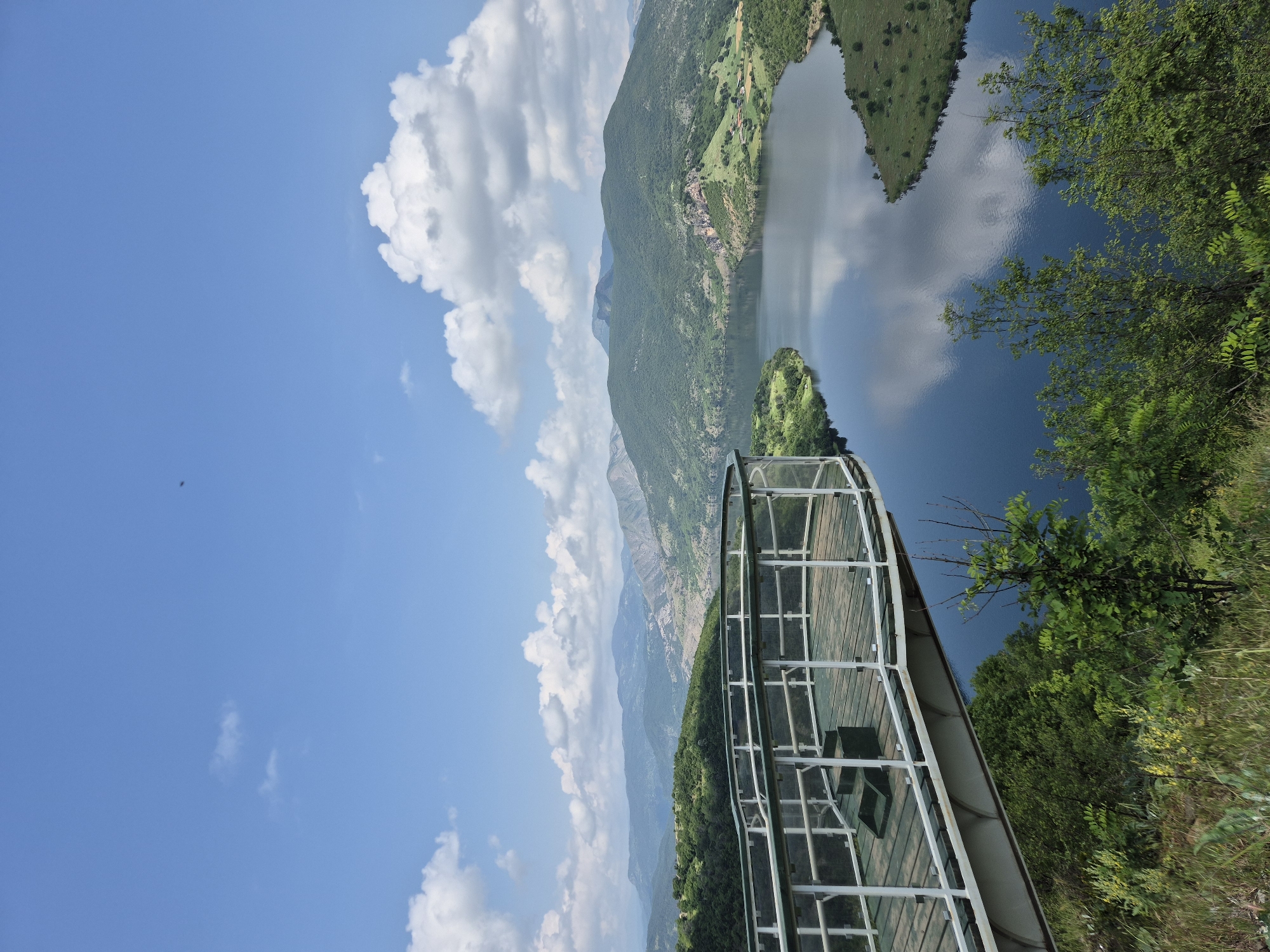 The image captures a panoramic view of a serene natural landscape from an elevated perspective in Ribartsi, Bulgaria. The central focus is a large body of water, likely a reservoir or lake, reflecting the bright blue sky and white, fluffy clouds above. This tranquil water is nestled amidst rolling, verdant mountains densely covered with trees and vegetation. In the far distance, additional mountain ranges can be seen under the expansive sky.
In the foreground, extending into the scene from the bottom right, is a modern viewing platform or walkway. It features a wooden deck surface and white or light-colored metal railings, possibly with glass panels, offering an unobstructed view of the scenery. Lush green trees and foliage frame the right side of the platform, enhancing the natural setting.
The scene depicts a bright, clear day, likely late morning or afternoon, indicated by the strong sunlight and well-defined shadows. The weather appears calm, with scattered cumulus clouds adding texture to the sky and reflecting gently on the water's surface. A tiny dark speck, possibly a bird or insect, is visible in the upper left sky. The overall impression is one of peace and natural beauty, inviting observation and contemplation. There is no visible text in the image.