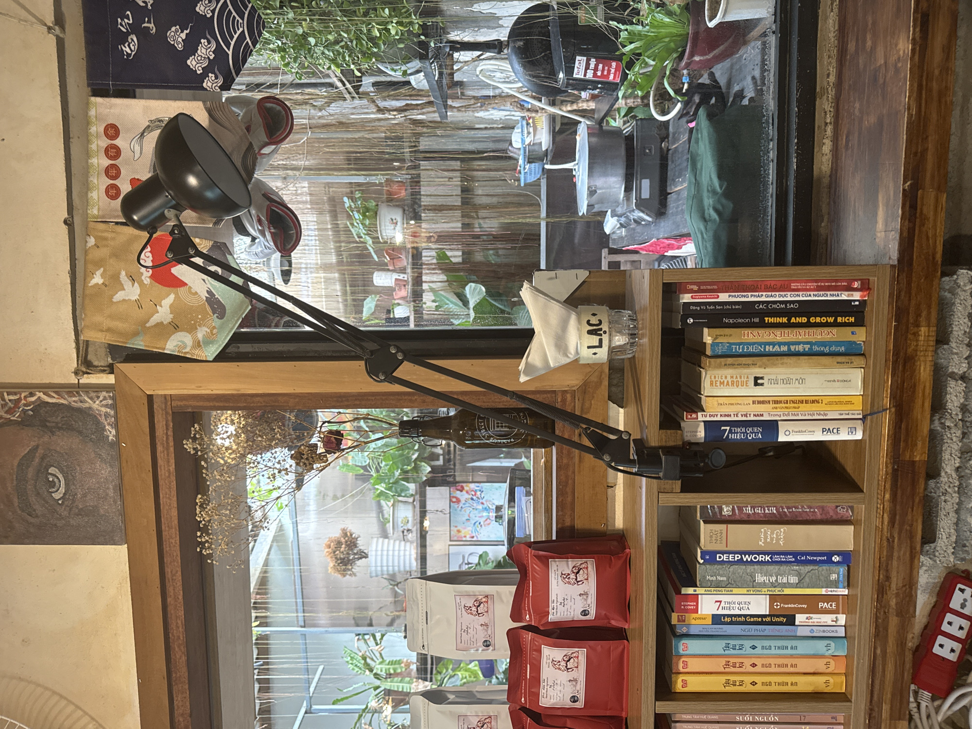 The image depicts an indoor scene, likely a cafe or a shop, with a prominent wooden shelf filled with books. The shelf is situated below a glass window that looks out onto a garden area with various plants. A black desk lamp with a flexible arm is positioned on the shelf, casting light. On the shelf, to the left of the lamp, are several bags of coffee. To the right of the lamp, there's a small stack of napkins in a glass holder with the word "LAC" written on it, and a metal pot on a portable stove in the background, suggesting food preparation. The overall ambiance is cozy and rustic, with natural light filtering through the window. The specific location in the city of Phú Thủy, Vietnam, is not directly indicated by any visual cues in the image.