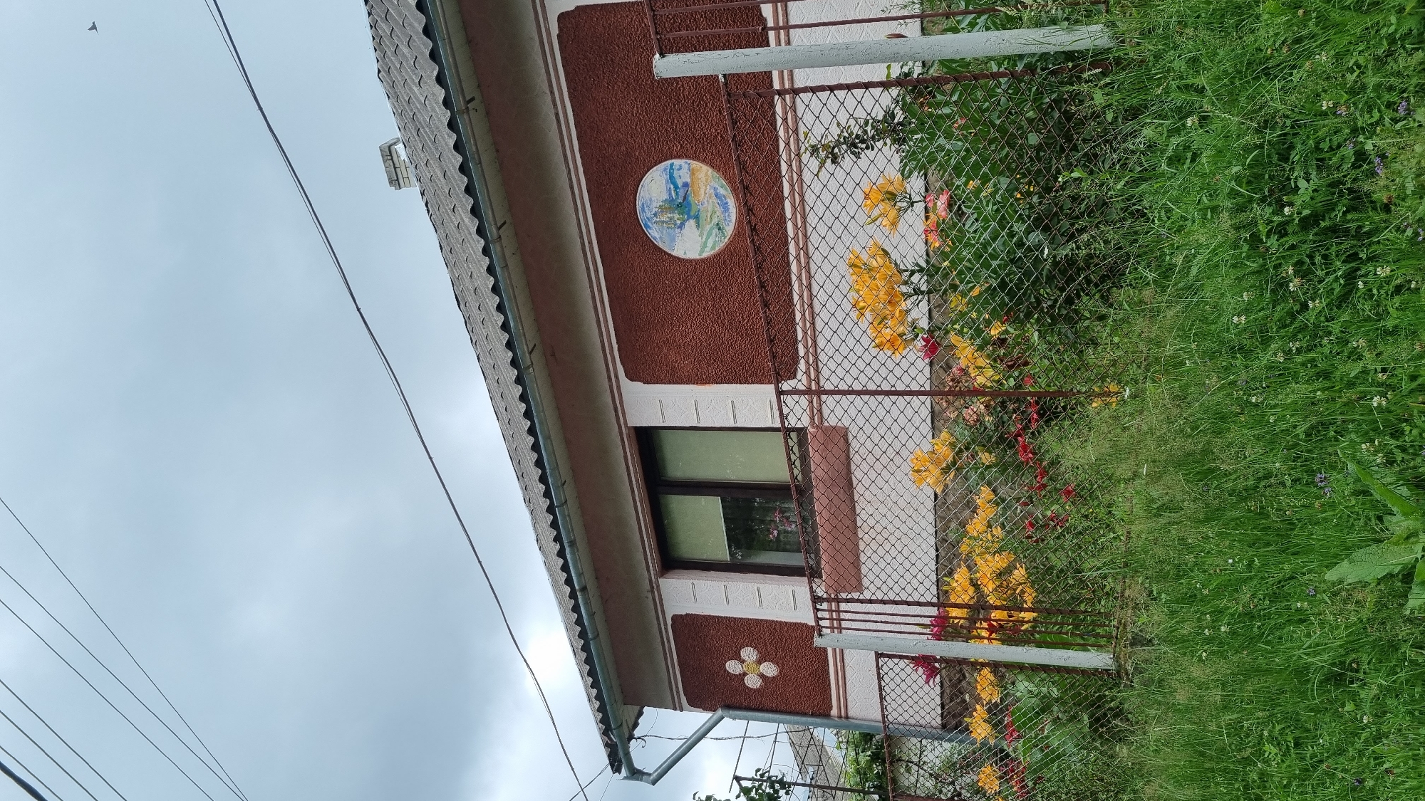 The image shows a section of a house with a brown textured facade and a pink trim. A circular decorative mosaic depicting a landscape scene is affixed to the facade above a window. Below the window, another decorative element resembling a four-petaled flower is visible. A chain-link fence runs along the front of the property, separating it from a lush green grassy area with wildflowers. Yellow and red flowers are blooming along the fence line, adding a splash of color to the scene. The sky is overcast, suggesting a cloudy day. Electrical wires are visible in the upper left portion of the image, and a bird is flying in the distance. The overall setting appears to be a residential area in Vyshka, Ukraine, likely during daytime. There is no visible text in the image.