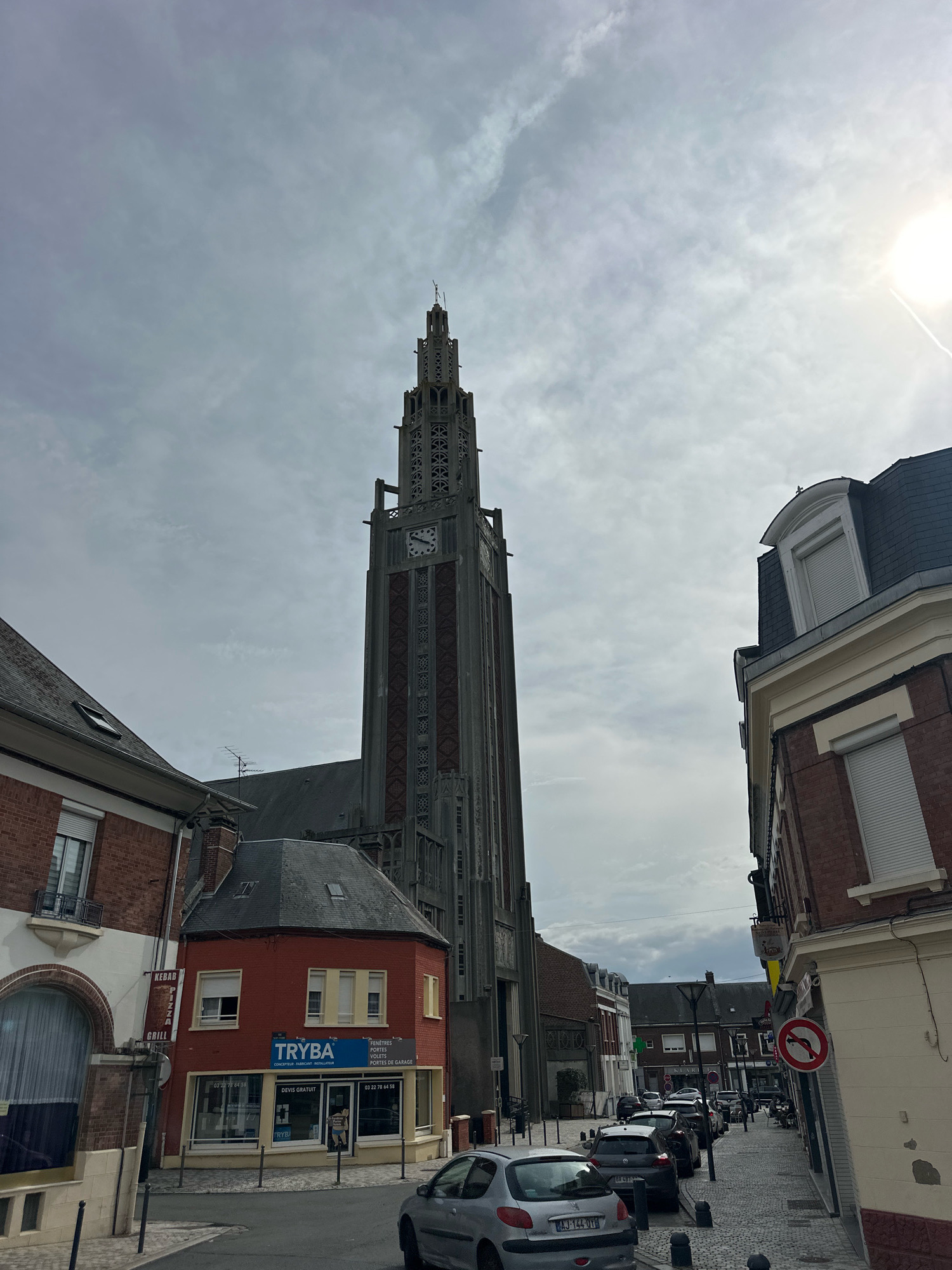 The image depicts a street scene in the city of Roye, France, dominated by the imposing tower of the Notre-Dame-de-la-Brèche church. The tower, constructed of concrete and brick, features intricate geometric patterns and a clock face. In the foreground, a red brick building houses a business named "TRYBA," which specializes in windows and doors, as indicated by signage. Several parked cars line the cobblestone street. To the right, another building with a mansard roof and brick facade is visible. The sky is overcast with the sun peeking through from the upper right corner, casting a slight glare. The scene appears to be set during the daytime.