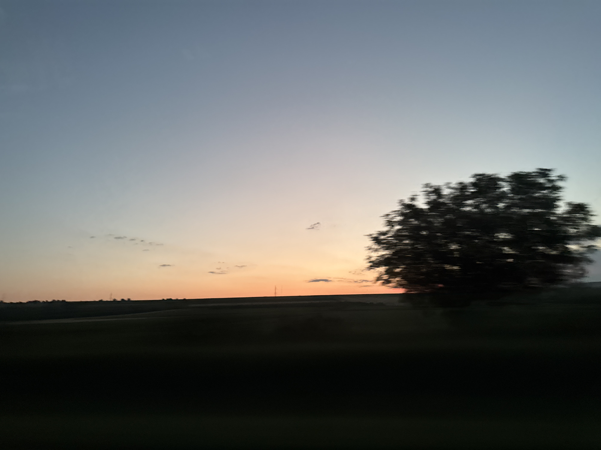 This is a photograph of a landscape at dusk. The sky is a gradient of pale blue at the top to soft orange and pink near the horizon. A dark, silhouetted tree is visible on the right side of the frame, blurred due to motion. In the distance, rolling hills are covered in dark green foliage, with a faint line of trees or structures along the horizon. A single, thin tower is also discernible on the horizon. The foreground is a dark, indistinct mass, likely representing fields or land. The overall impression is of a rural scene captured from a moving vehicle.