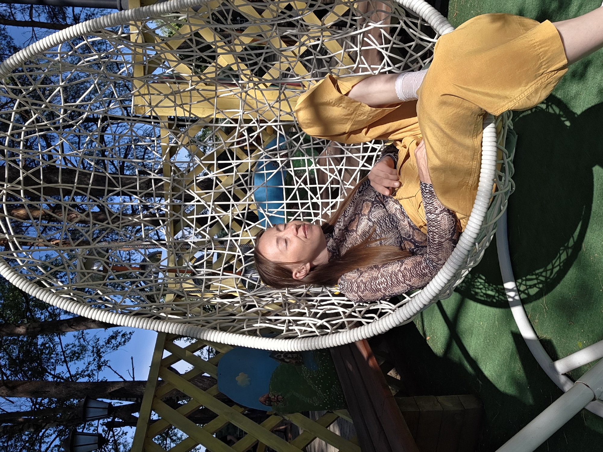 A woman is reclining in a white hanging wicker chair in what appears to be an outdoor setting. She is wearing a brown snakeskin-print long-sleeved shirt and mustard-yellow pants. Her left ankle is wrapped in what looks like a bandage or light-colored fabric. The chair is partially shaded, casting a shadow on a green surface below. Through the wicker, trees and blue sky are visible. A yellow lattice structure with painted details is present in the background. The location is Chaikovsky, Russia.