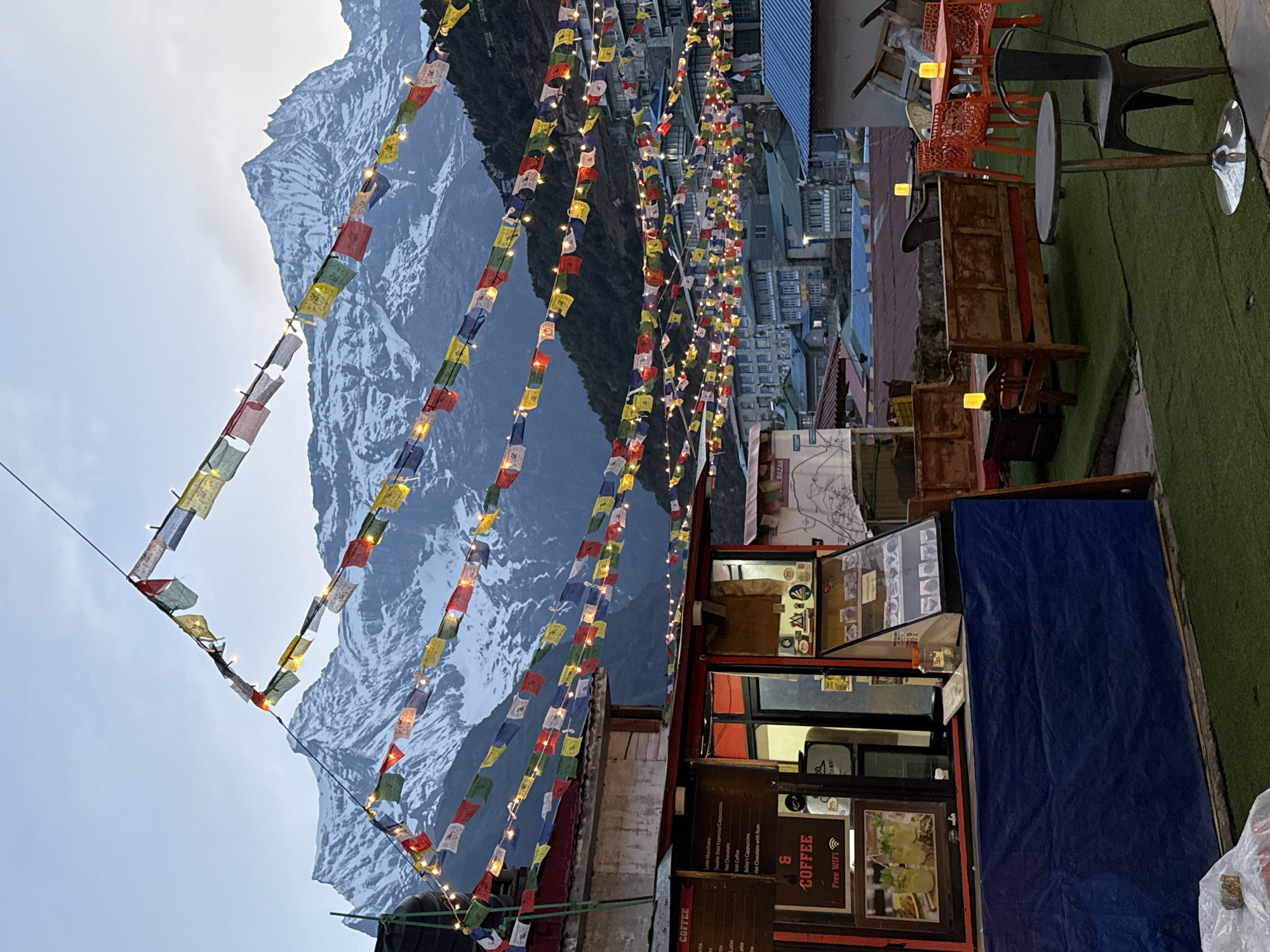 The image captures a vibrant outdoor café or resting area nestled in the mountainous landscape of Namche, Nepal, likely during dusk or an overcast day. In the background, majestic snow-capped peaks rise dramatically under a soft, diffused sky.
Numerous colorful Tibetan prayer flags, strung with small twinkling lights, crisscross the scene, creating a festive and spiritual canopy overhead. These flags are a prominent visual element, guiding the eye from the foreground towards the formidable mountains.
In the mid-ground and foreground, a cozy outdoor seating area is visible. To the right, a small coffee shop or stall with red trim and windows offers refreshments. Its sign prominently displays "COFFEE" and a menu including "Latte Macchiato," "Double Shot Espresso/Cappuccino," "Chocolate," "Bailey's Cappuccino," and "Hot Chocolate with Rum," along with "Free WIFI." A partially visible word "BAZAR" can also be seen.
Adjacent to the coffee stall, the seating area features artificial green turf on the ground, holding various furniture pieces. These include bright orange plastic chairs arranged around tables, a tall bar-style table with a single dark chair, and rustic wooden benches. Small yellow lanterns or candles illuminate some of the tables, adding to the warm and inviting ambiance. In the distance, other buildings of the Namche settlement can be glimpsed, blending into the rugged terrain.
The scene conveys a sense of serene beauty and comfort, where trekkers or visitors can enjoy warm beverages and relaxation amidst the breathtaking Himalayan scenery, enhanced by the twinkling lights and traditional prayer flags.