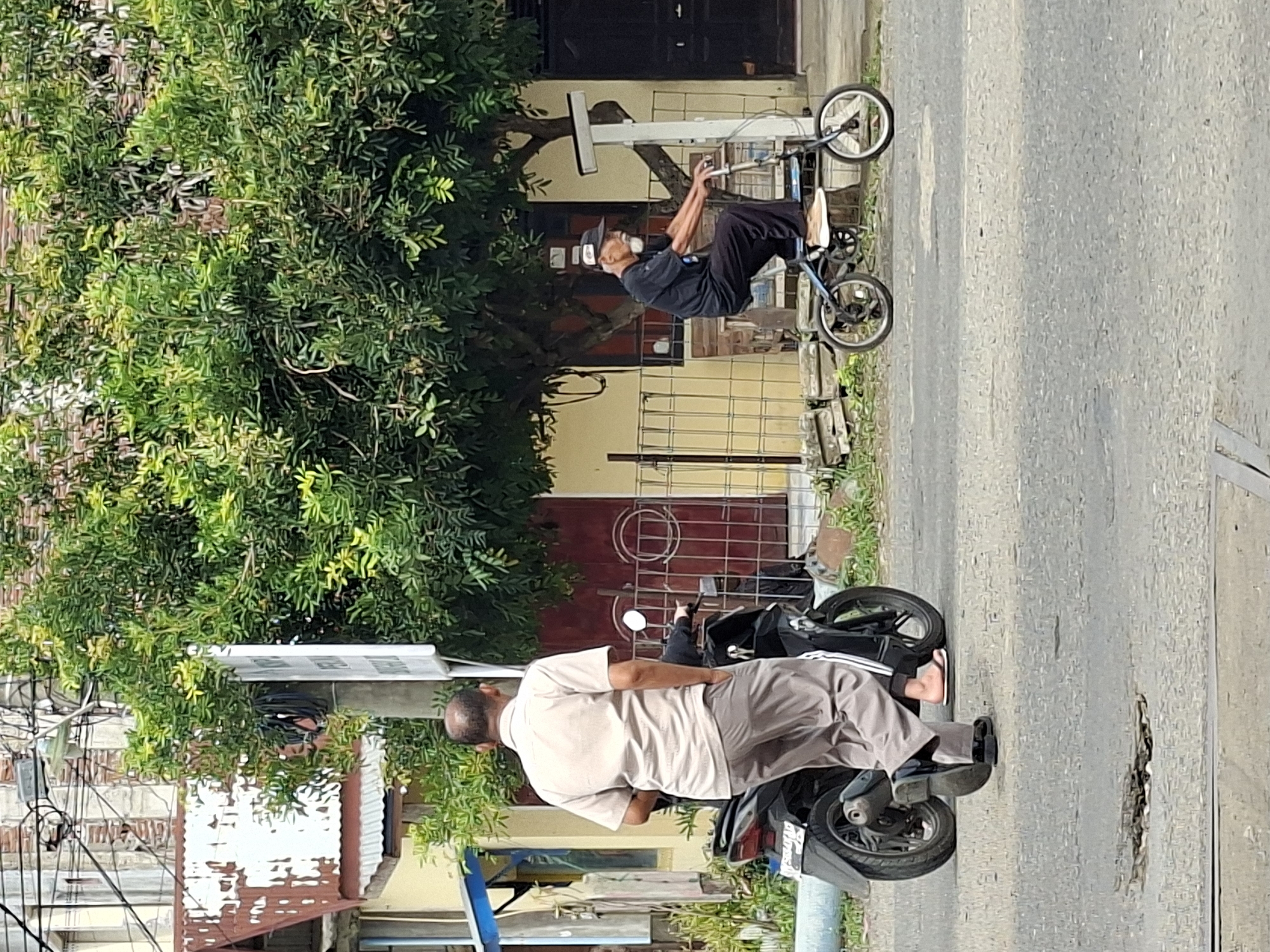In Lhokseumawe, Indonesia, an elderly man wearing a black t-shirt, black pants, and a baseball cap rides a small blue foldable bicycle on a paved street. He has a white beard and mustache and is looking forward. To his left, a man wearing a light brown collared shirt and grey pants stands next to a black scooter. His right hand is in his pocket, and his left arm is bent. The scooter is parked on the side of the road. In the background, a yellow building with red doors and a corrugated metal roof is visible. Lush green trees and foliage surround the area, with a metal fence in front of the building. The street has a pothole in the foreground, and utility poles and wires are present overhead.