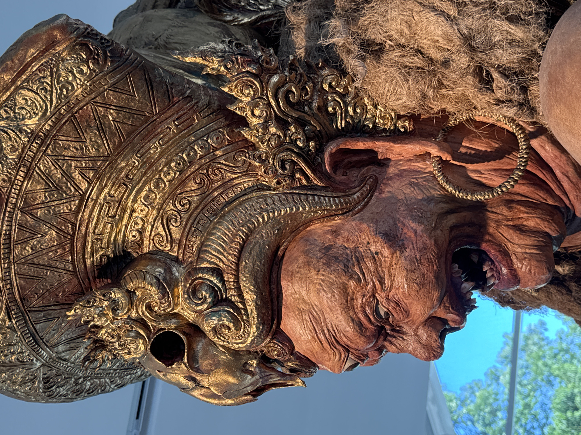 This is a close-up, eye-level, medium shot of a large, carved sculpture depicting a grotesque, demon-like figure. The figure's head is adorned with an elaborate, golden headdress featuring intricate patterns and what appears to be a skull motif. The sculpture's skin is a reddish-brown, with deeply wrinkled features, bulging eyes, and an open mouth revealing sharp teeth. A large, golden hoop earring is visible on the figure's ear, and a mass of coarse, brown hair surrounds its head. The background is out of focus, showing a bright blue sky with some green foliage visible, suggesting an outdoor setting or a backdrop. The lighting is strong, casting shadows that highlight the texture of the sculpture. The sculpture is located in Kuta Selatan, Indonesia, likely as part of a cultural display or performance art.