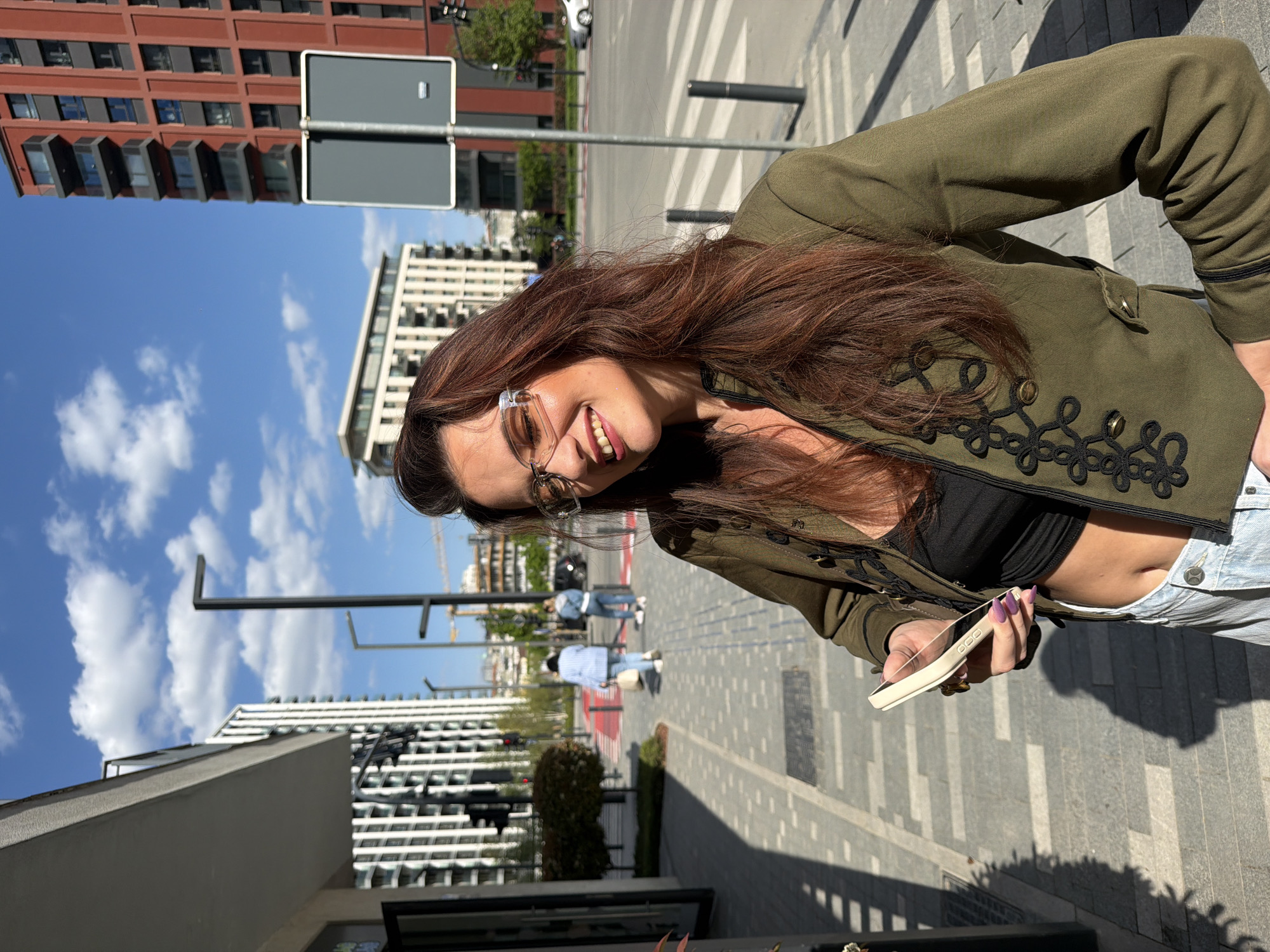 The image, presented rotated 90 degrees counter-clockwise, captures a woman smiling broadly at the camera in an urban environment, likely Belgrade, Serbia. The main subject, with long, wavy brown hair, wears clear-framed glasses, an olive green military-style jacket with black embroidered details and gold buttons, a black crop top, and light blue jeans. Her midriff is exposed. She holds a white smartphone in her right hand, revealing purple nail polish and a ring on her finger. Her expression is joyful and relaxed.
The background shows a clear blue sky with scattered white clouds, indicating a bright, sunny day, likely late morning or early afternoon, given the long shadows on the pavement. The urban setting includes a prominent red-brick building with numerous windows on the left and modern white and grey buildings further back on the right. Paved sidewalks with a patterned design lead to a road with a zebra crossing. Several blurred figures are visible walking in the distance, including one in a light blue top and jeans, and another in white. Street signs, street lamps, and a partially visible construction crane add to the city landscape. No visible text is discernible in the image.