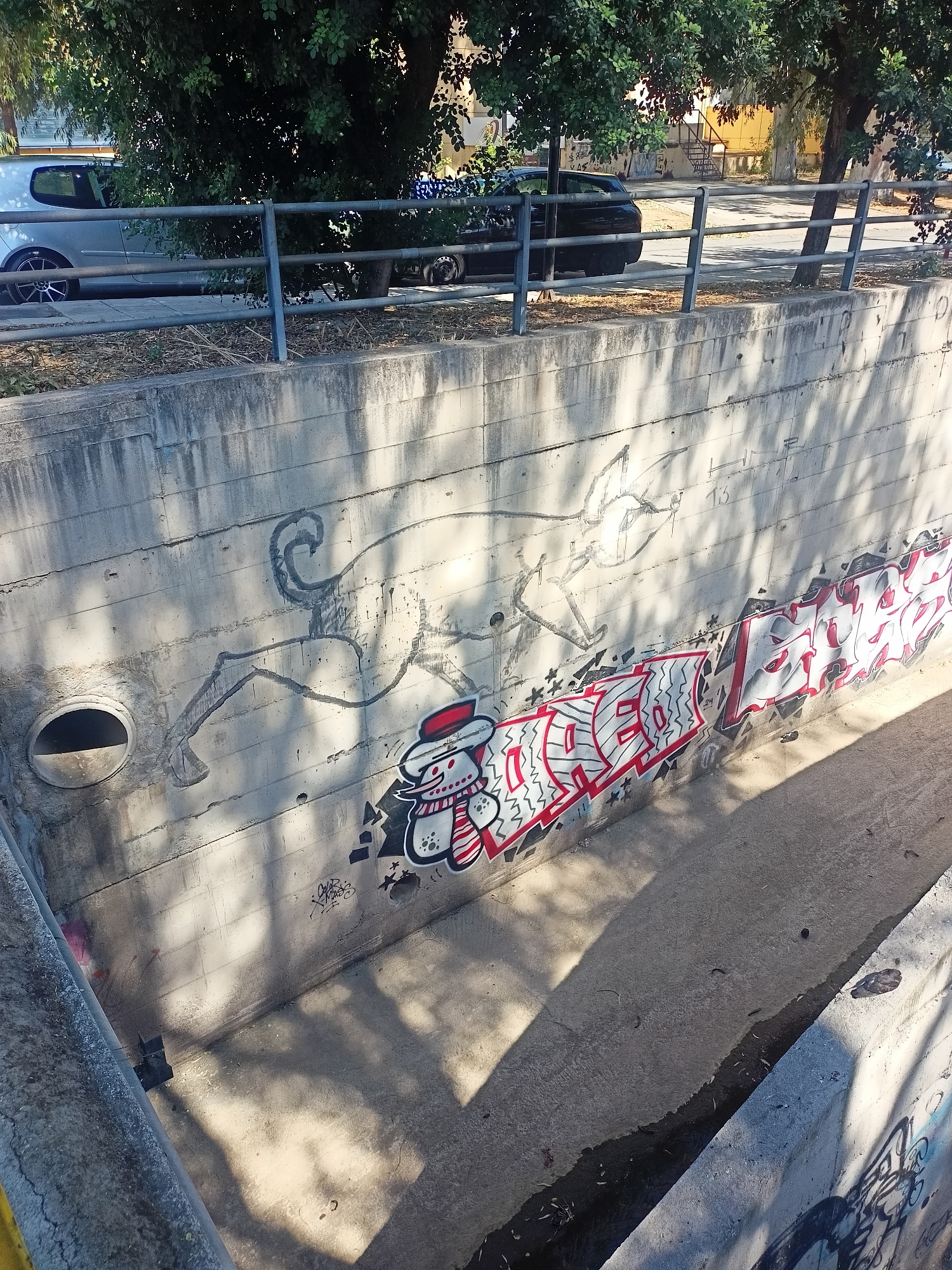 An outdoor, medium-shot, high-angle photograph captures a concrete drainage channel adorned with graffiti art under dappled sunlight. On the left side of the channel's wall, a large, stylized drawing of a dog or wolf-like creature with a long, flowing tail and a pointed muzzle is rendered in black. To the right of this drawing, a snowman character with a red hat and scarf is depicted, followed by a section of graffiti with bold, blocky letters in red and white. The graffiti extends further to the right, showing more red and white lettering with black outlines and shadows. The concrete wall has a rough, textured surface with visible horizontal lines from the concrete forms. Shadows of trees and foliage are cast across the wall and the channel floor, suggesting an urban park or roadside setting. A metal railing runs along the top edge of the channel on the left, and on the right side, part of another concrete wall with graffiti is visible. Cars are parked on a street above the drainage channel, partially obscured by trees.