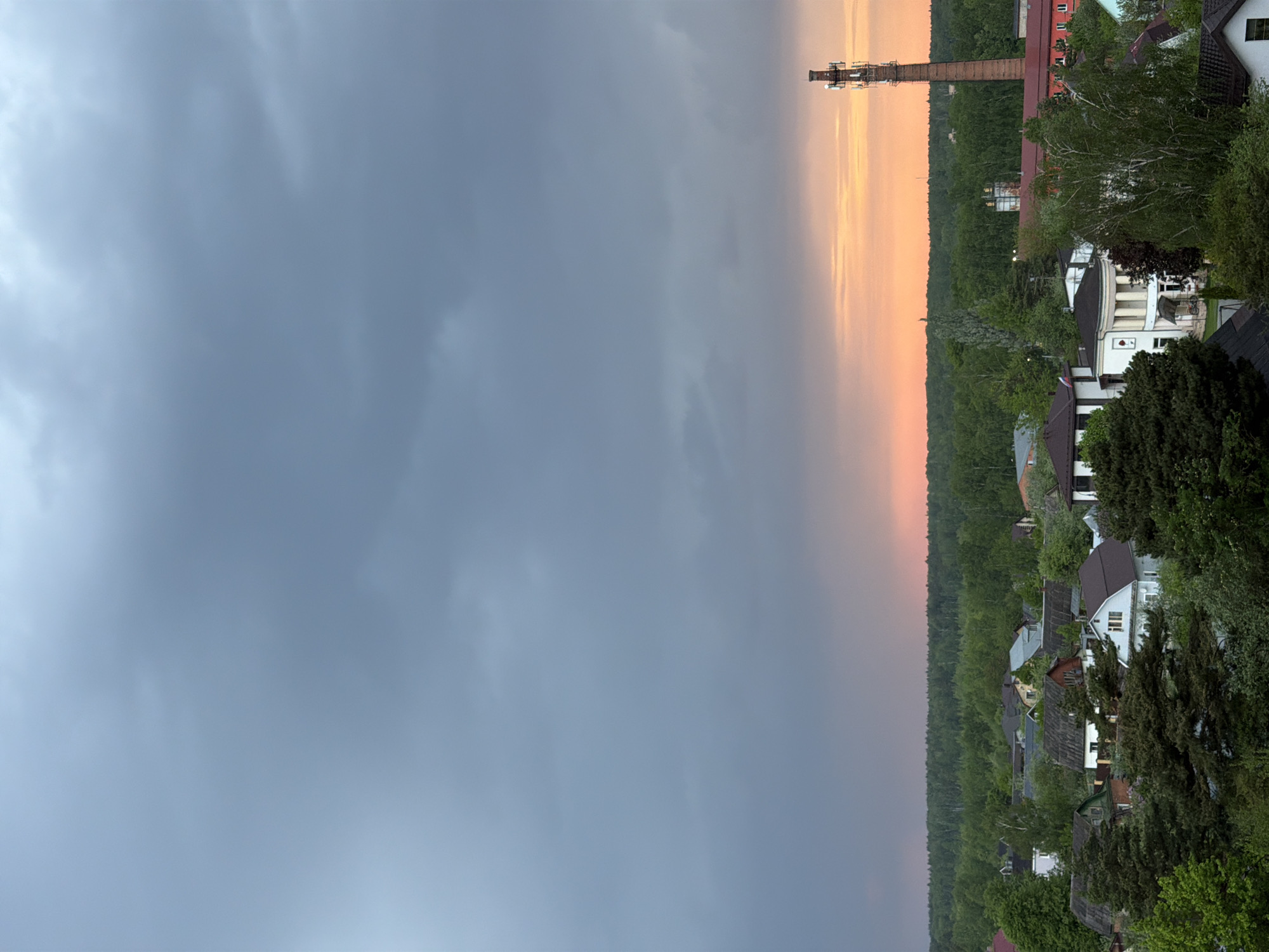 The image shows a landscape with a gray, cloudy sky occupying most of the frame. A vertical gradient transitions to a soft orange hue on the right, where a tall brick chimney stands next to a structure with scaffolding. Lush greenery and trees fill the middle ground, interspersed with houses of varying sizes and architectural styles. The scene is likely located in a suburban or rural area within a city.