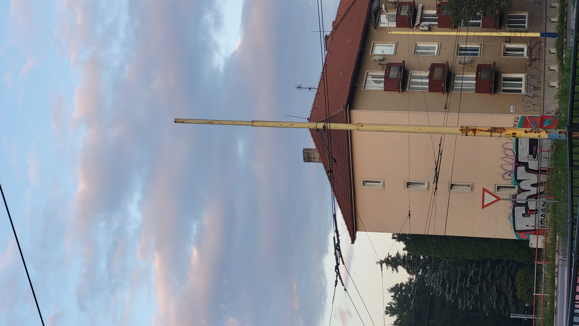 The image shows a building in the city of Prešov, Slovakia. The building is a multi-story apartment complex with a red tiled roof. The exterior walls are a light beige color. There are several windows and balconies visible. The lower part of the building is covered in graffiti. In front of the building, there is a traffic light pole and overhead power lines. Trees are visible in the background, and the sky is partly cloudy with hints of pink and orange, suggesting it might be sunset. There are no people visible in the image.
