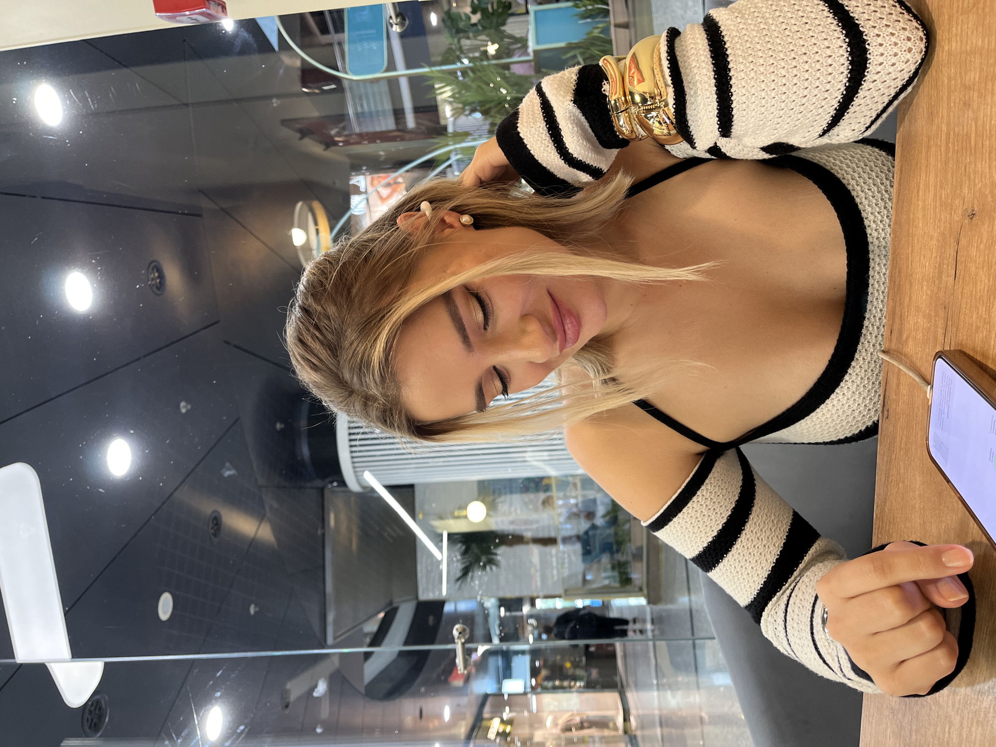 A young Caucasian woman with blonde hair is seated at a wooden table, looking down at her smartphone. She is wearing a black and white striped top with detached sleeves. Her right hand is resting on the table, pointing towards the phone, while her left hand is touching her hair. She is wearing a pearl earring and a large gold bracelet on her right arm. The background suggests an indoor location, possibly a cafe or shopping mall, with glass partitions, interior lighting, and other patrons visible. There is no specific indication of the city being Moscow, Russia.