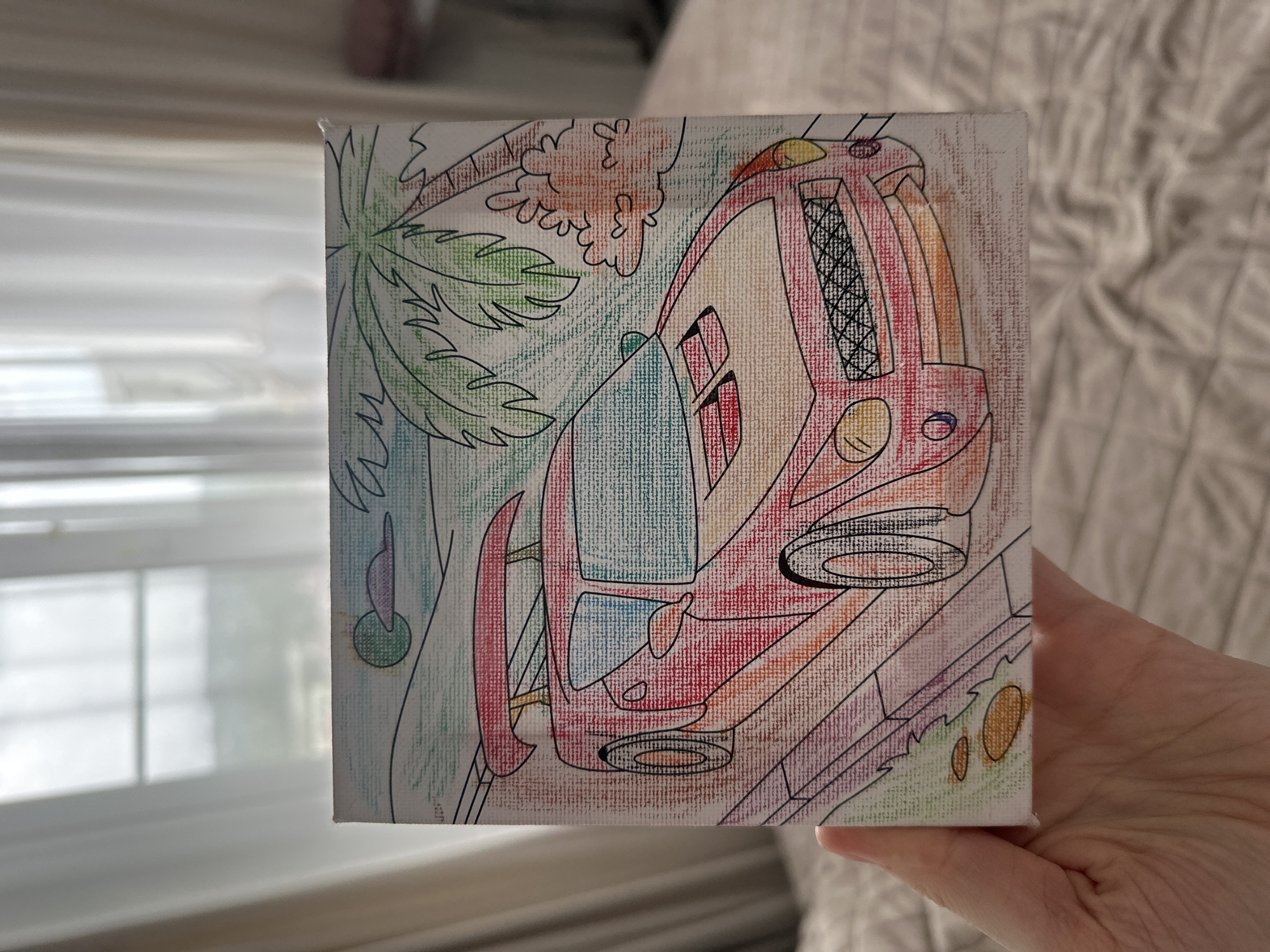 A square canvas painting held by a person's hand depicts a stylized, futuristic car driving along a road. The car is predominantly red, with details in orange, yellow, and black. It features an unusual design, including a rounded front, a prominent grill, and distinct wheels. The backdrop includes a green palm tree and other vague foliage, suggesting a tropical or coastal environment. The scene is rendered with visible pencil strokes and a simplistic, childlike aesthetic. The location in Sochi, Russia, is not evident from the painting itself.