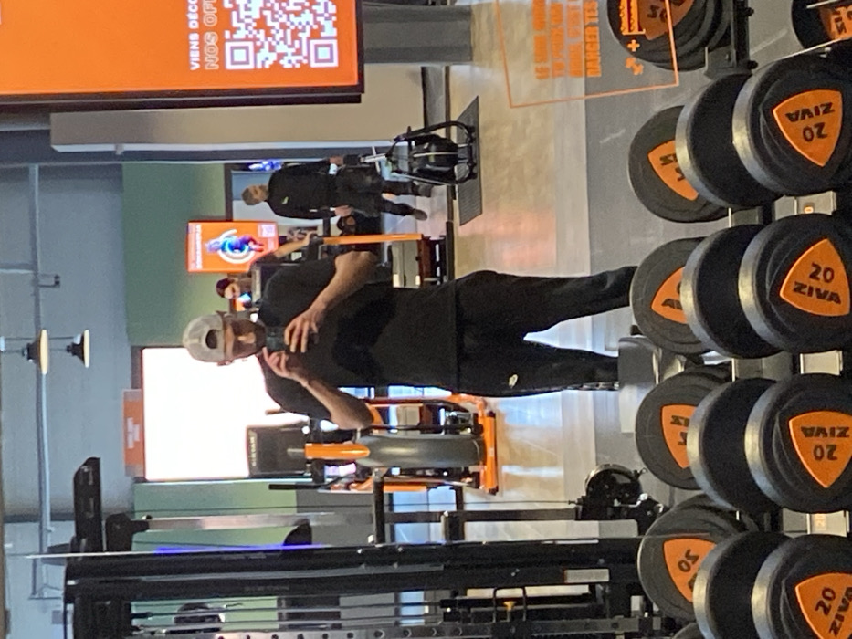 The image depicts a reflection in a large mirror within a fitness gym, specifically located in Hœnheim, France. In the foreground, a rack holds numerous dumbbells with black hexagonal ends and orange triangular inserts, marked with "ZIVA" and weight values, such as "20 ZIVA".
The primary subject, a male individual in a mid-shot reflection, stands centered. He wears a grey baseball cap backward, a black short-sleeved t-shirt, and black sweatpants with a white Nike logo visible on the left thigh. He has a beard and is holding a dark-colored smartphone in front of his chest, looking down at the device, presumably engaging with it. A thin bracelet is visible on his left wrist.
In the reflected background, to the left of the primary subject, a black cable machine and other exercise equipment with orange accents are visible. To the right of the primary subject, another individual, possibly female with dark hair, is partially visible, appearing to be on a cardio machine. Further in the background, a third male individual in a dark long-sleeved shirt and dark pants is walking towards the right, near a black rowing machine positioned on a black mat.
A prominent vertical orange screen on the right displays French text, including "VIENS DÉCOUVRIR NOS OFFRES!" (Come discover our offers!) and a QR code. Another smaller orange sign with an abstract blue and orange figure is visible in the distant background. The gym features light-colored walls, a light floor, and industrial-style dome ceiling lights.