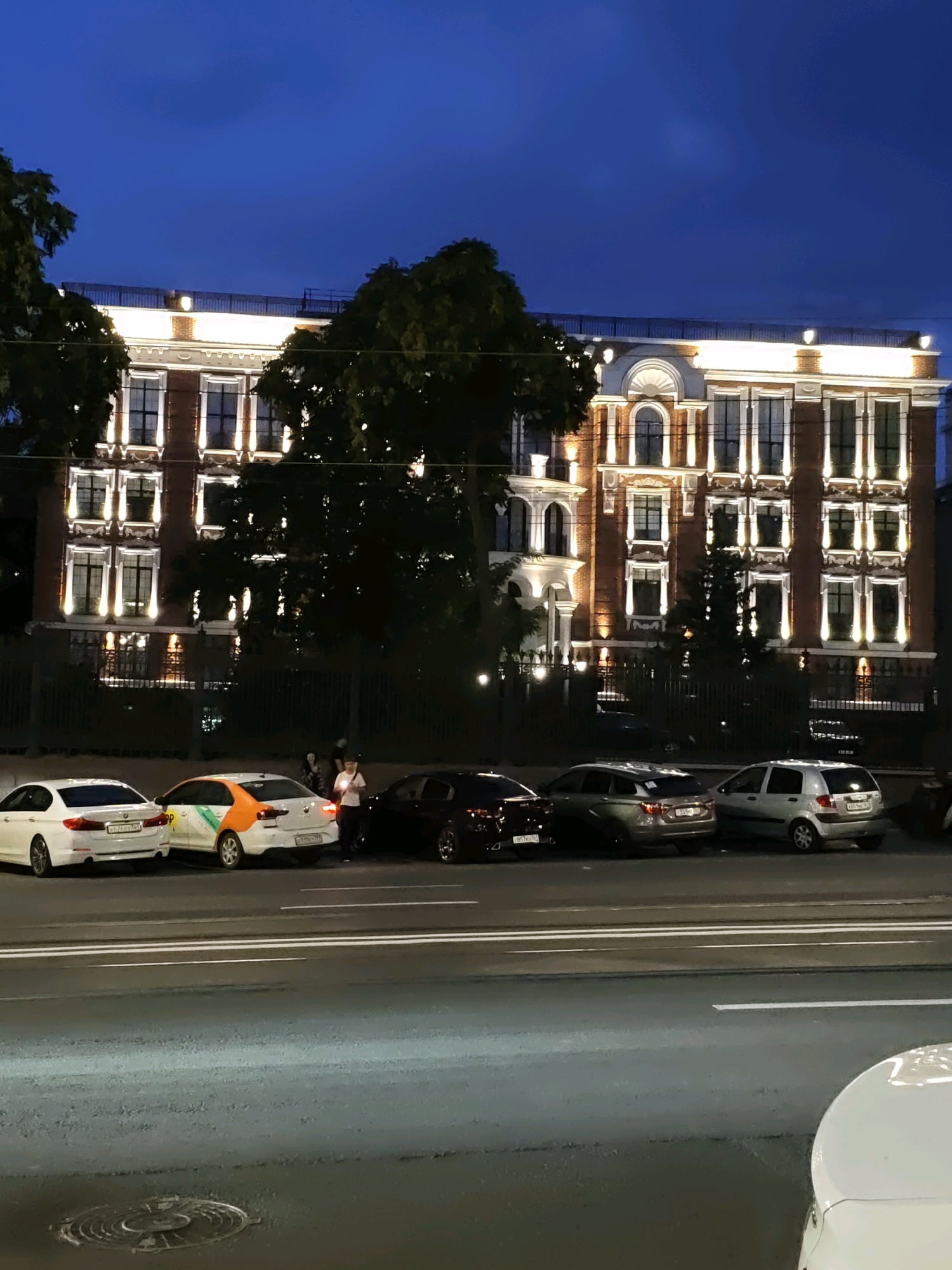 A nighttime exterior shot captures a grand, illuminated building in Rostov-on-Don, Russia. The architecture features multiple stories with rows of windows, each outlined by bright lights that create a striking contrast against the deep blue twilight sky. The building appears to be made of brick with white decorative elements. In front of the building, a street is visible with several cars parked along the side. One car is white, another is orange and white, and there are dark-colored vehicles as well. A few people are standing near the cars, engaged in conversation or activity. Trees with dark foliage partially obscure the building and the street. The scene is lit by the building's exterior lights and the ambient light of the evening, casting long shadows and creating a slightly dramatic atmosphere.