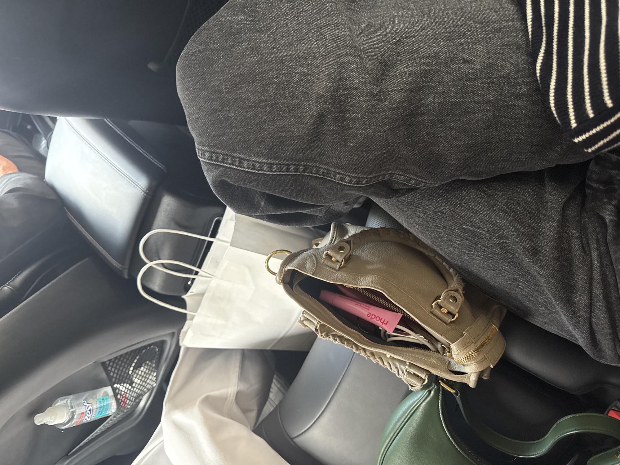 The image captures an interior scene within a vehicle, likely a car or taxi, observed from an overhead perspective. A person's lower body is visible, dressed in dark grey denim jeans and a black and white striped knit top, occupying a dark-colored seat.
Prominently featured in the foreground is a light taupe or khaki-colored leather handbag with gold-toned hardware, sitting open on the person's lap. Inside the open bag, a pink tube labeled "rhode" is clearly visible. To the bottom right, a portion of a dark green leather crescent-shaped handbag is also seen.
To the left of the person's legs, two white paper shopping bags with handles are present. On the car's dark door panel, a mesh pocket holds a small, clear spray bottle of hand sanitizer, partially labeled "Zest." The car's interior appears to have dark leather seating and dark panels.
The scene takes place during the daytime, illuminated by soft, ambient light that creates subtle reflections on the car's surfaces. The activity depicted is passive, suggesting the person is seated, possibly traveling or waiting, in Mexico City, Mexico. No specific emotions are discernible.
