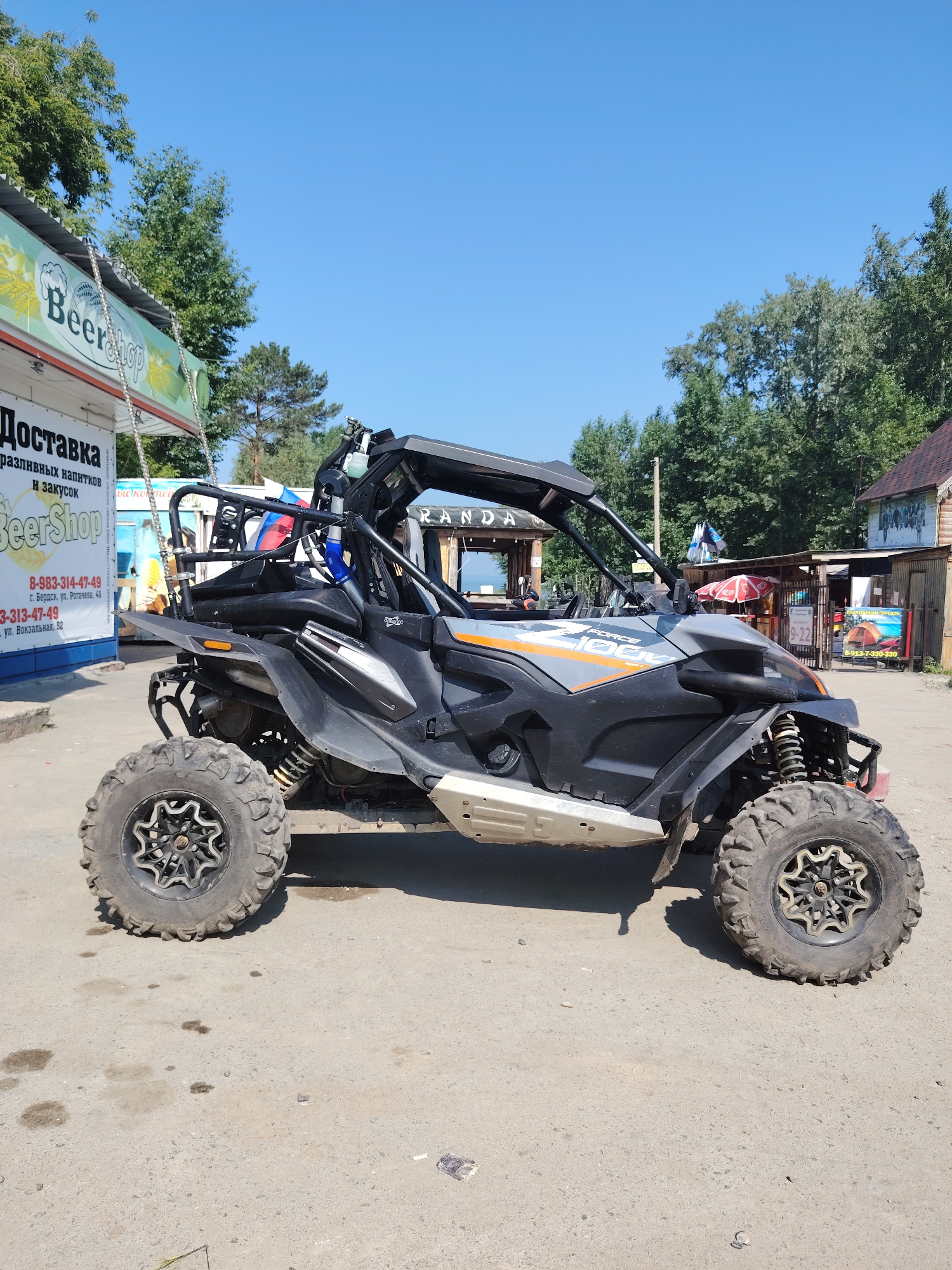 A side-view, medium shot captures a black and grey off-road vehicle parked on a dusty, cracked asphalt surface under a bright, clear blue sky. The vehicle, a CFMOTO Force 1000, is positioned slightly angled towards the viewer's left. Its prominent off-road tires with distinctive silver rims are visible, as are its suspension components.
Behind the vehicle, a small, open-air establishment is present. A sign on the left side of the establishment reads "BeerShop" and "Доставка разливных напитков и закусок" (Delivery of draft beverages and snacks). Another sign further back displays "RANDA". There are also wooden structures and fences, with some trees visible in the background. Two small red and white umbrellas are set up near one of the structures. A Russian flag is attached to a metal bar on the back of the off-road vehicle. The scene appears to be a roadside or recreational area.