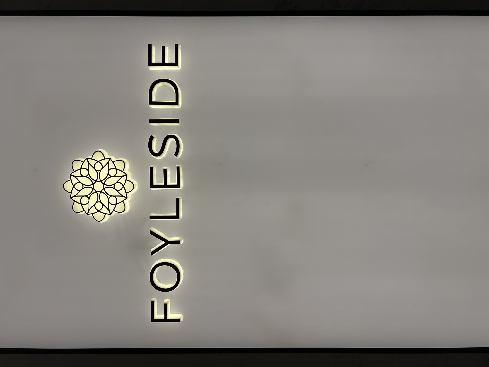 The image displays illuminated signage on a light-colored wall. Above the text "FOYLESIDE," there is a circular logo with a floral or mandala-like design, also illuminated. The "FOYLESIDE" text is in a sans-serif font with a black outline and a yellow-white glow emanating from behind the letters. The signage appears to be installed indoors, possibly in a mall or commercial building, given the clean, minimalist background. The city of Derry/Londonderry is known for the Foyleside Shopping Centre, a prominent retail hub. Therefore, this signage is likely located at the Foyleside Shopping Centre in Derry/Londonderry, United Kingdom. No people are visible in the image.