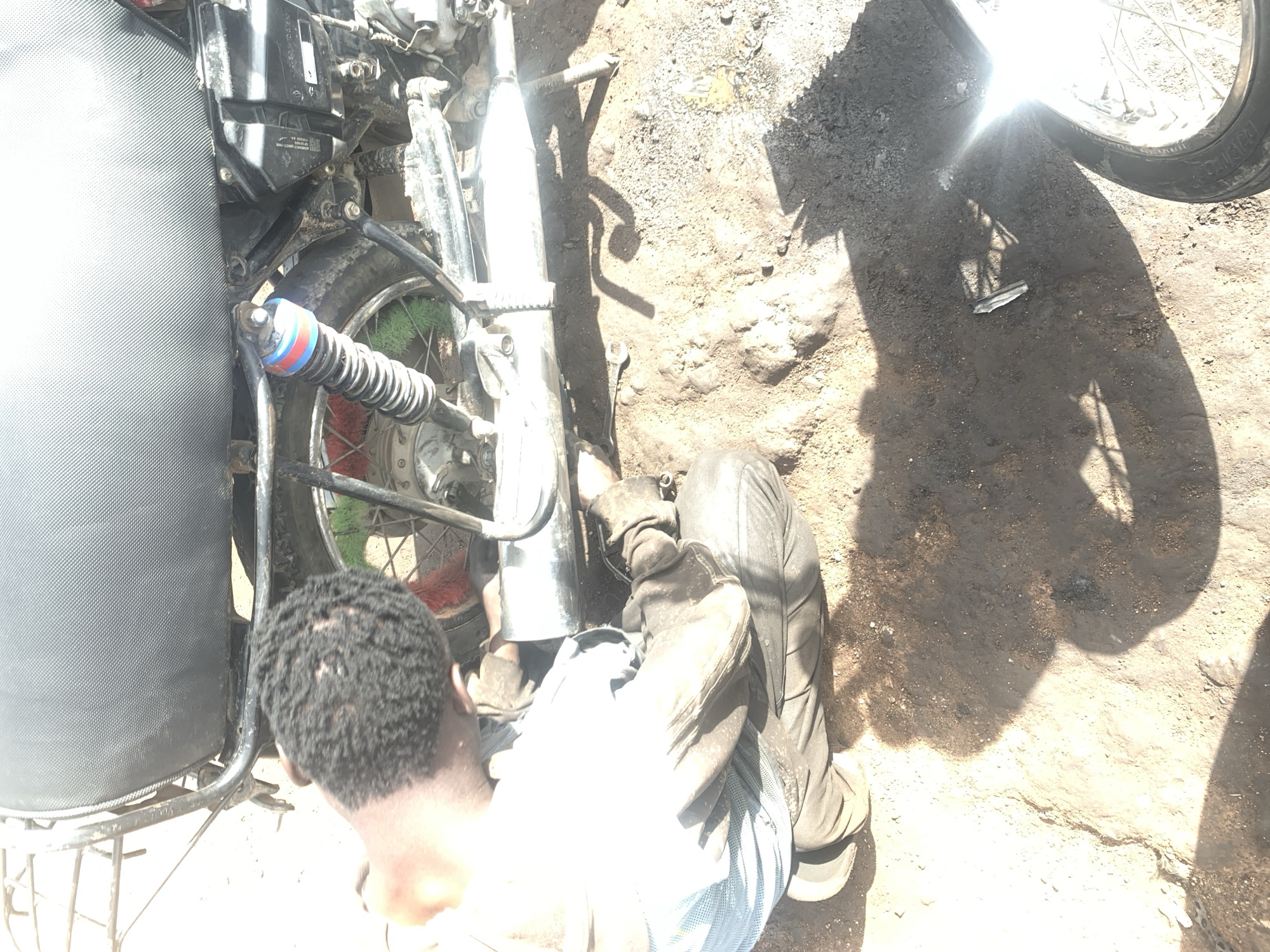 In Gura Top, Nigeria, a person is seen working on a motorcycle. The individual, with braided dark hair, is wearing a light-colored long-sleeved shirt and dark pants. They are crouched down, appearing to be engaged in some repair or maintenance of the motorcycle's rear wheel and exhaust system. A shock absorber with red and blue bands is visible, as well as parts of the motorcycle's frame and seat. The ground is covered in dirt and debris, suggesting an outdoor or workshop setting. Sunlight casts strong shadows, indicating it is daytime. A wrench is visible on the ground near the motorcycle.