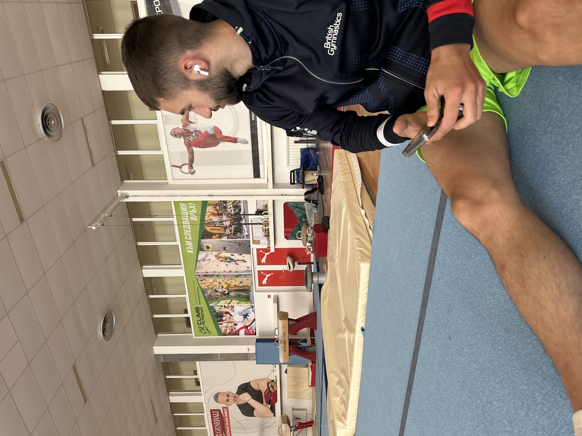 The image captures a man sitting in what appears to be a gymnastics training facility, possibly in Sofia, Bulgaria given the Bulgarian text on the walls. He's wearing a dark blue British Gymnastics jacket with a red and white accent on the sleeve and bright green shorts. He is wearing a wireless earbud in his left ear. He is holding a smartphone in his hands, examining it.
The setting includes gymnastics equipment such as a pommel horse and a balance beam. Walls are decorated with large promotional posters featuring images of gymnasts and advertisements for a climbing academy and a Generali insurance company; one poster has the Bulgarian text "КЪМ СЛЕДВАЩИЯ ВРЪХ!", which translates to "To the next peak!". The facility floor is a light blue padded mat. The lighting seems to be fluorescent, typical for indoor gym environments.