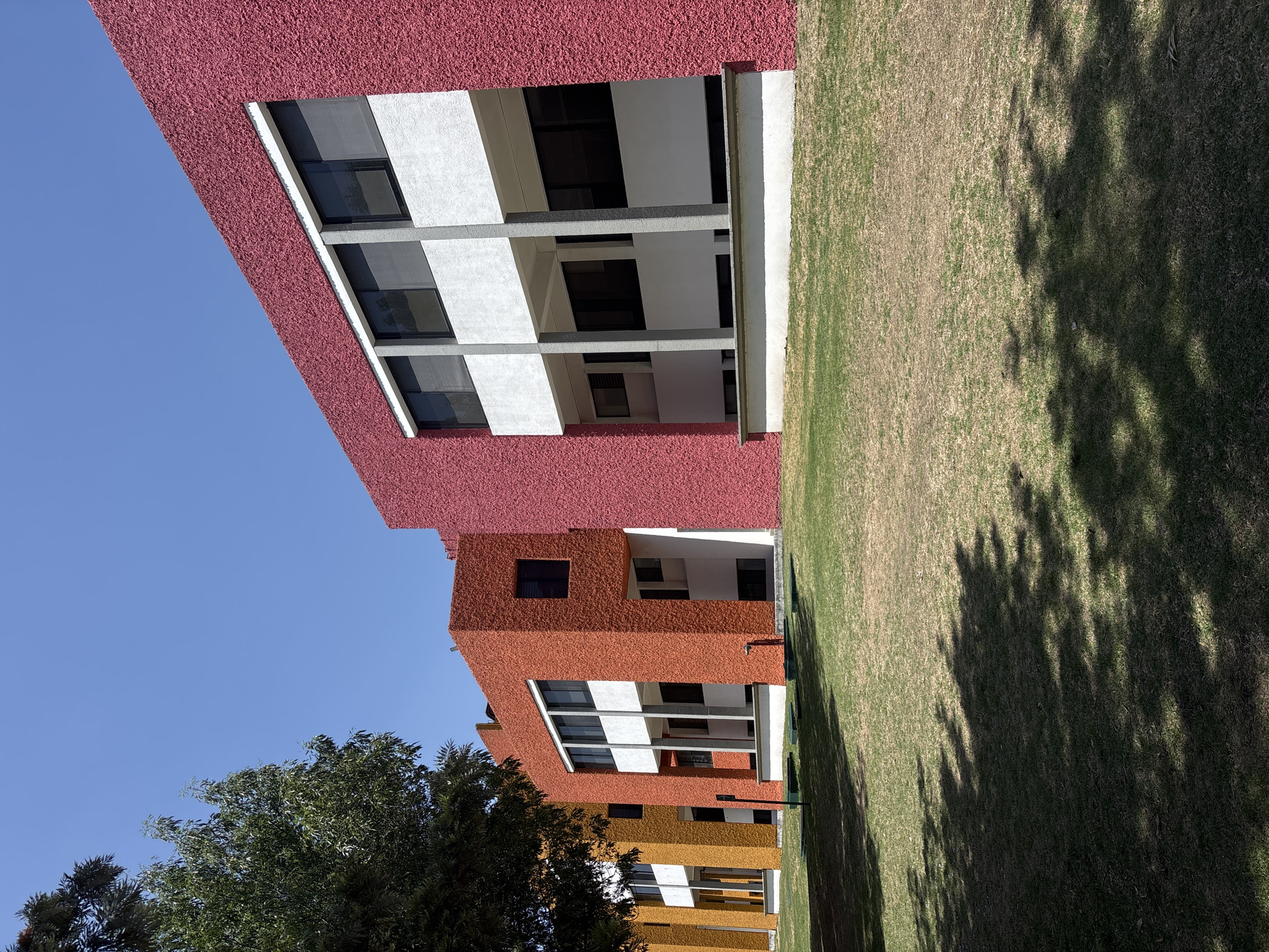 The photograph captures a row of multi-story buildings situated alongside a grassy area. The buildings exhibit a modern architectural style characterized by their cubic shapes and a color palette transitioning from red at the top to orange and yellow in the lower sections. The facades feature textured surfaces, and rectangular windows are interspersed throughout. The grassy area occupies a significant portion of the right side of the image and is partially covered in shadows cast by nearby trees. The sky above is clear and blue, indicating a sunny day. The location is San Andrés Cholula, Mexico.