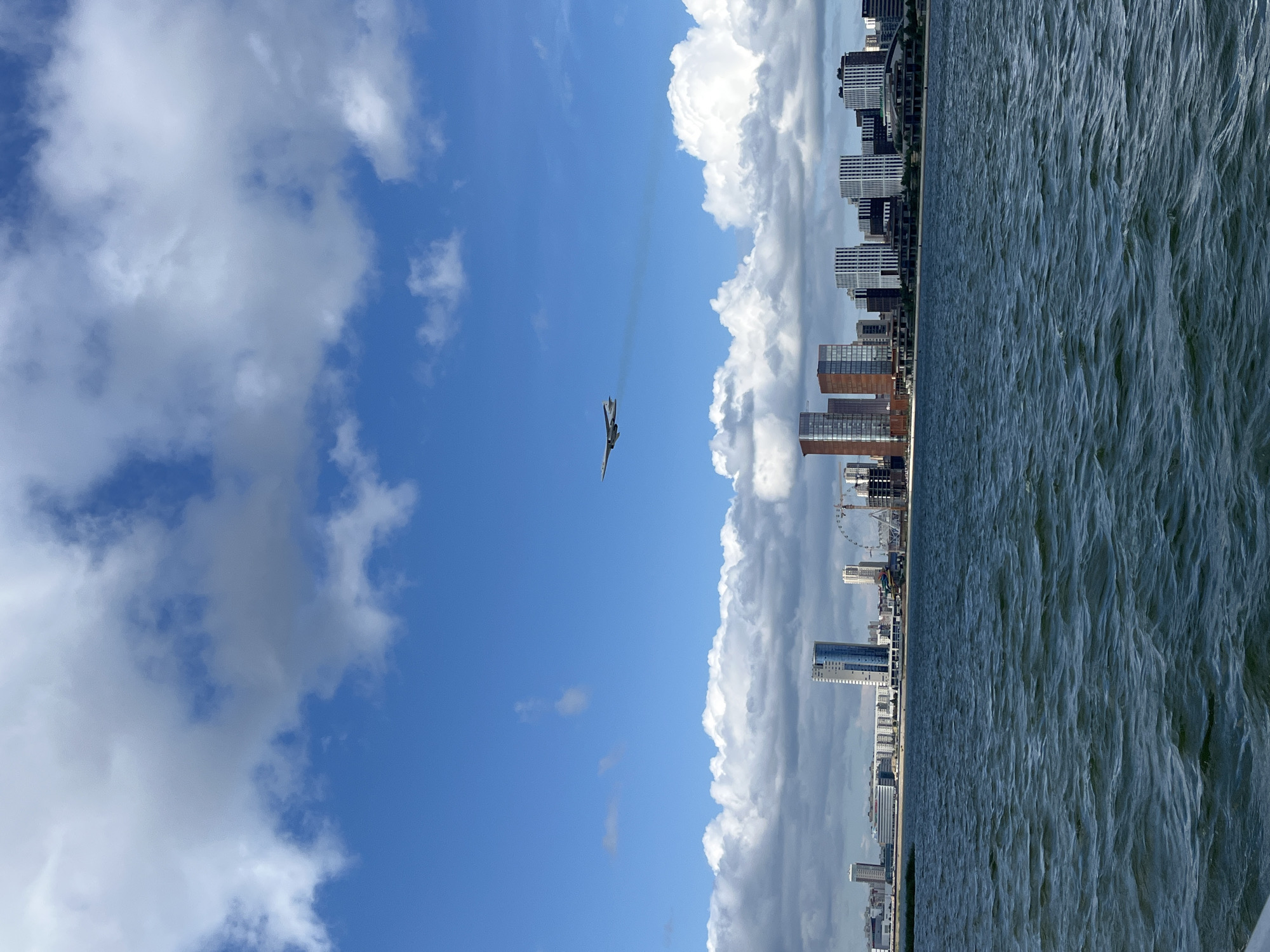 A jet airplane flies through a bright blue sky with scattered white clouds, leaving a faint contrail behind it. Below the airplane, a city skyline rises from the shore, featuring a mix of modern skyscrapers and other buildings. The buildings appear to be clustered together, with some taller structures standing out among them. A large Ferris wheel is visible in the distance, indicating a potential entertainment or recreational area. The foreground is dominated by a body of water, likely a river or lake, with visible ripples on its surface, suggesting a slight breeze. The overall scene depicts an urban waterfront with aerial activity. This location is the city of Kazan, Russia, likely along the Kazanka River or the Kuybyshev Reservoir.