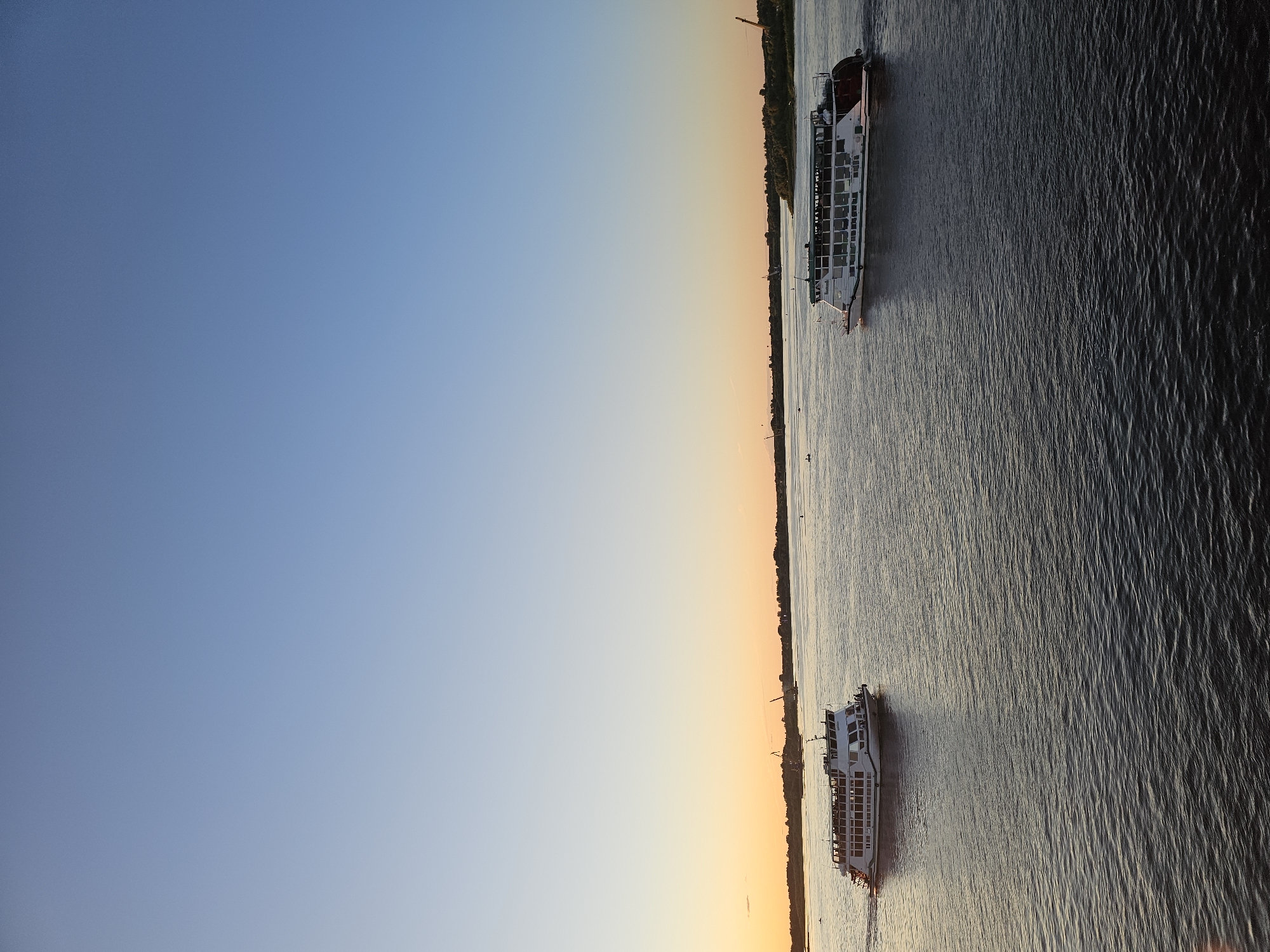 The image shows a panoramic view of a wide river at sunset, likely in Nizhny Novgorod, Russia. The sky transitions from a clear, deep blue on the left to a warm gradient of orange and yellow near the horizon on the right, indicating the time of day is evening. The water surface is rippled, reflecting the colors of the sky and the light from the setting sun.
Two passenger boats are visible on the river, moving in the same direction. The larger boat, positioned further away and towards the top right of the frame, is white with multiple decks and rows of windows. It appears to be a cruise ship or a large tour boat. The second, smaller boat is closer to the bottom right, also white with windows, suggesting it is also a passenger vessel. The boats are leaving a slight wake behind them, indicating movement.
Along the far bank of the river, a line of trees and some structures are discernible, forming a distinct horizon. A crane is visible on the left side of the distant shore. The overall mood of the image is serene and tranquil, amplified by the soft lighting and the vastness of the river and sky. There is no visible text in the image.