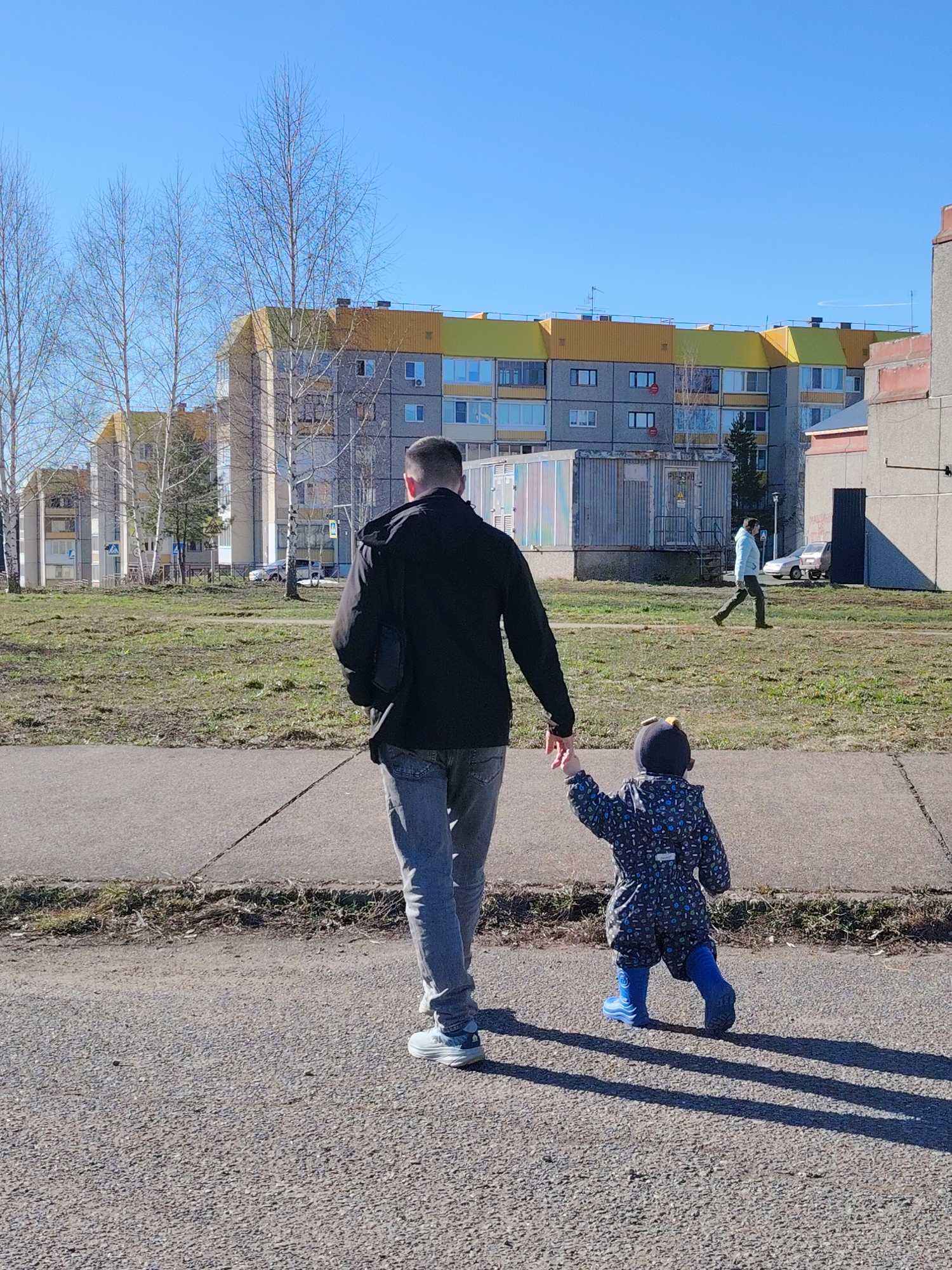 The image depicts a man and a small child walking away from the camera, holding hands, on an asphalt path under a clear blue sky. The man, seen from behind, wears a black jacket, grey jeans, and light-colored sneakers, carrying a black shoulder bag over his right shoulder. The child, also viewed from the back, is dressed in a dark blue patterned full-body suit, bright blue rubber boots, and a dark hat.
The setting is an outdoor urban environment, consistent with a residential area in Марковский, Russia. In the background, several multi-story apartment buildings are visible, characterized by grey facades and distinctive yellow and light green roof sections. The surrounding landscape consists of sparse, dry grass and numerous leafless deciduous trees, indicating a late winter or early spring season. Long, distinct shadows of the man and child are cast on the asphalt, suggesting bright sunshine and a low sun angle, likely mid-morning or late afternoon.
Further in the background, another person walks across a grassy area, and a light-colored car is partially visible near one of the buildings. A utilitarian-looking container structure stands between the foreground and the apartment blocks. While some signs or notices are visible on building windows, their text is unreadable. The overall scene portrays a calm, everyday moment of a guardian and child walking together.