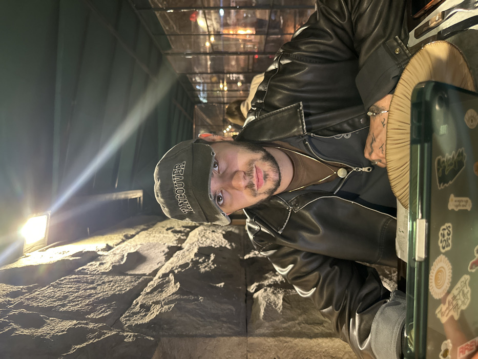 The image depicts an indoor or semi-outdoor restaurant setting at night in Yerevan, Armenia. A male subject, appearing to be in his 20s or 30s, is seated at a table, looking directly at the camera. He has short dark hair, a beard, and a mustache. He is wearing a distressed dark grey or olive green baseball cap with "VANCOUVER" embroidered in white, a dark brown leather jacket, and a brown t-shirt. Two gold-colored necklaces are visible, one with a circular pendant and another with a cylindrical pendant. A dark-colored tattoo is visible on his left hand. His forearms rest on a dark table, beneath which a white menu or placemat is partially visible on the left. A dark green smartphone, adorned with multiple stickers, is positioned in the foreground on the left. To his right, a light-colored, possibly wooden, fluted plate is present, with silverware (knife and fork) visible further to the right. The background features a textured, dark stone wall on the left, illuminated by a bright overhead light fixture, which casts a prominent lens flare. To the right of the subject, a dark-colored overhead structure or ceiling is visible, along with translucent partition panels with vertical framing. Out of focus in the background, another individual with light hair and a light-colored top is seated, facing away from the camera. Ambient lights are blurred in the distant background.