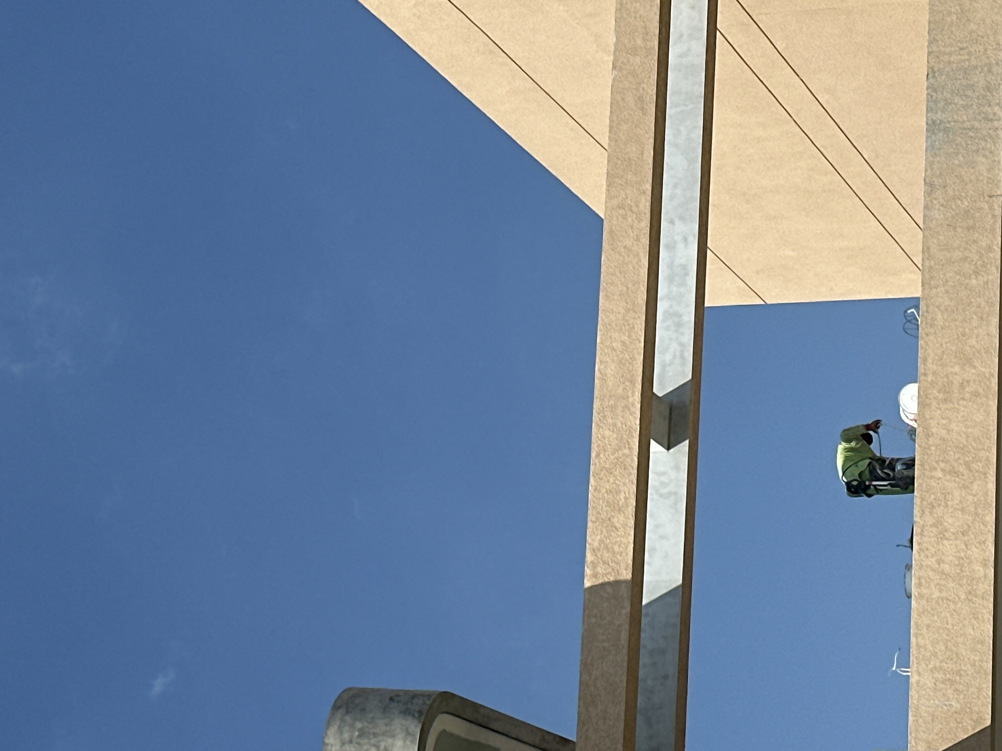 In the city of North Kuta, Indonesia, a construction worker wearing a bright green vest and dark pants is bent over on a beige concrete ledge. The worker appears to be engaged in some sort of construction or maintenance activity, with tools and a white bucket visible nearby. The sky is a clear, vibrant blue, with a few wispy clouds. The scene is framed by architectural elements, including a large concrete pillar on the left and a beige angled wall with diagonal lines on the right. A horizontal beige beam with a metallic underside runs across the middle of the image, providing a sense of depth and structure to the scene. The image captures a moment of labor against a bright, open sky.