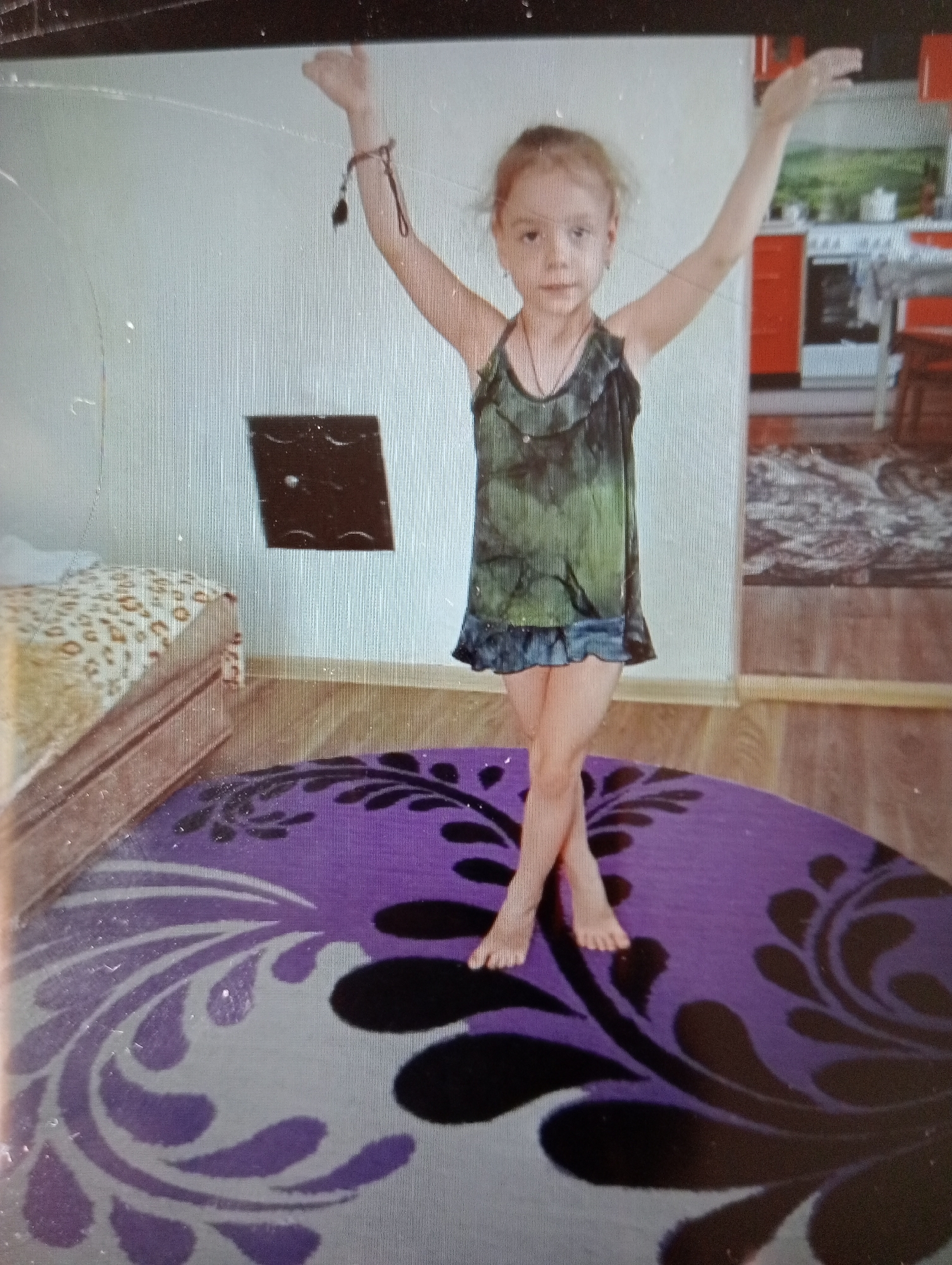 A young girl stands on a purple rug with a floral pattern. She is wearing a green and black tie-dye tank top and a denim skirt. Her arms are raised in the air, and her legs are crossed. She has blonde hair and is looking at the camera with a neutral expression.
In the background, there is a bed with a leopard-print comforter, a brown wooden floor, and a wall with a dark, square object hanging on it. Further in the background, a kitchen area is visible, with red cabinets and a white stove.
The lighting suggests it is daytime, possibly indoors in a home. There is no visible text in the image.