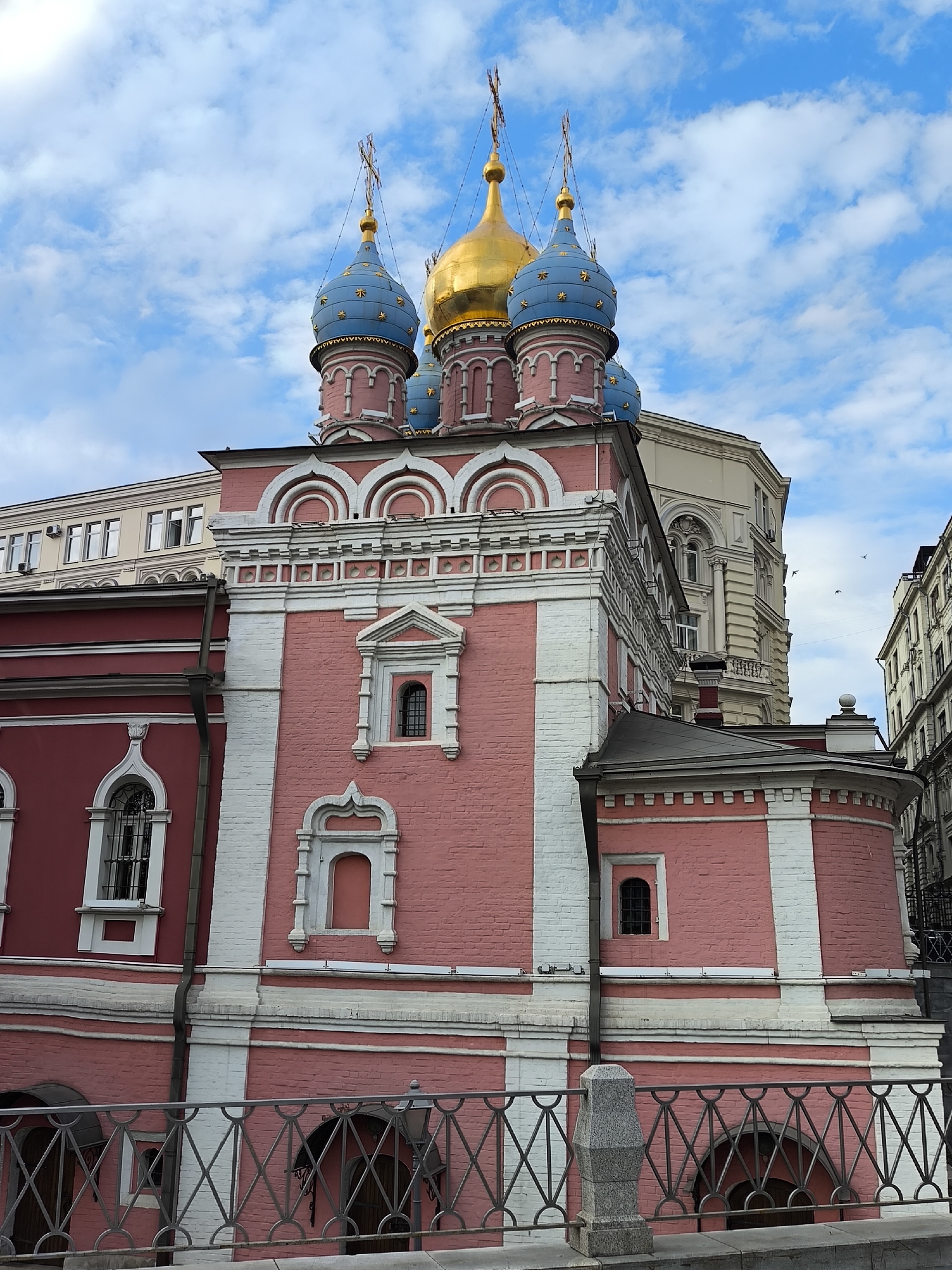 The image depicts a Russian Orthodox church, characterized by its pink brick facade with white accents. The church is topped with a golden central dome and smaller blue domes, each adorned with a golden cross. The sky is partly cloudy. The church is bordered by a wrought-iron fence with concrete posts. The building is in an urban setting in the city of Unknown, Unknown. Adjacent buildings are visible, suggesting a dense urban environment.