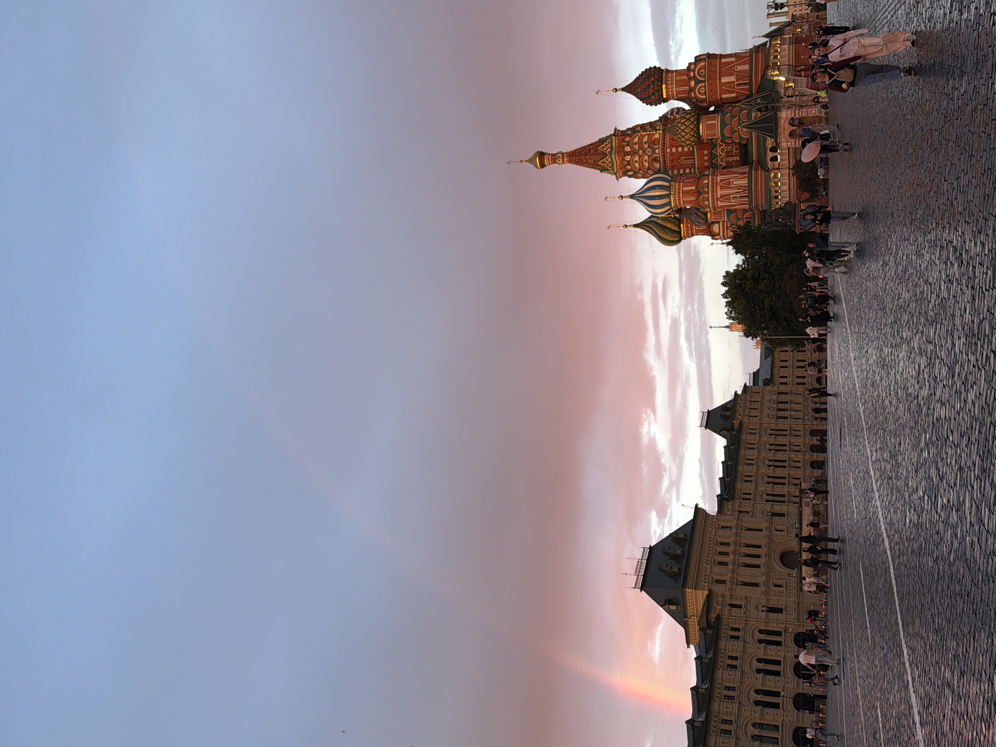 The image depicts Red Square in Moscow, Russia, at dusk. A faint rainbow is visible in the sky, arching over the scene. On the left, the GUM department store, a prominent landmark, stands with its tan facade and arched windows. On the right, Saint Basil's Cathedral with its iconic colorful onion domes is prominently featured. The ground is paved with cobblestones. Several people are walking across the square, some in small groups, engaging in casual activity. The sky is a gradient of blue and pink hues, indicating the setting sun.