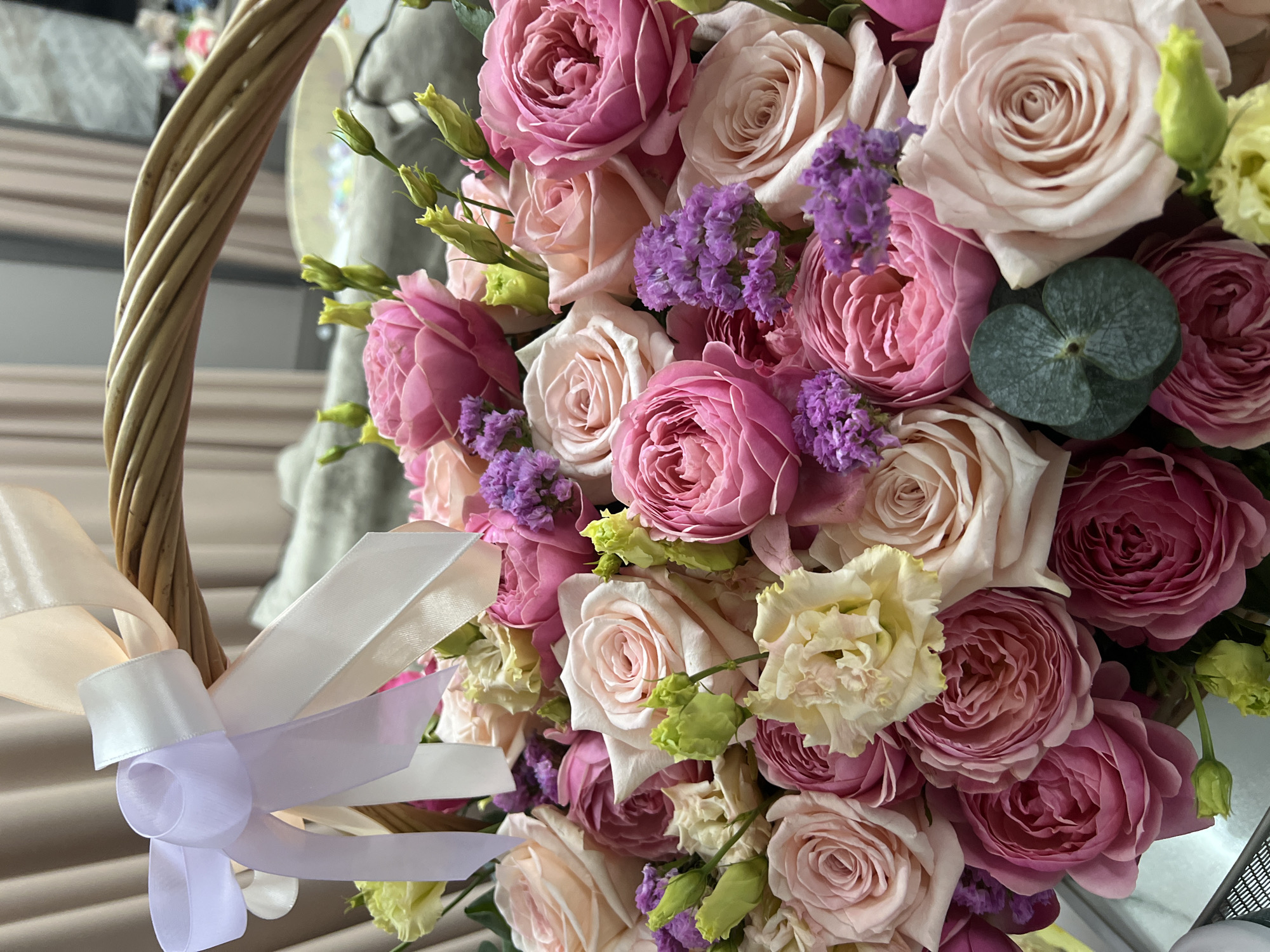 A close-up shot showcases a floral arrangement within a woven basket. The flowers primarily consist of roses in shades of pink and pale peach, interspersed with small clusters of purple blossoms. A white satin ribbon adorns the basket handle, tied in a bow. The background is blurred, suggesting an interior setting, potentially in Tyumen, Russia.
