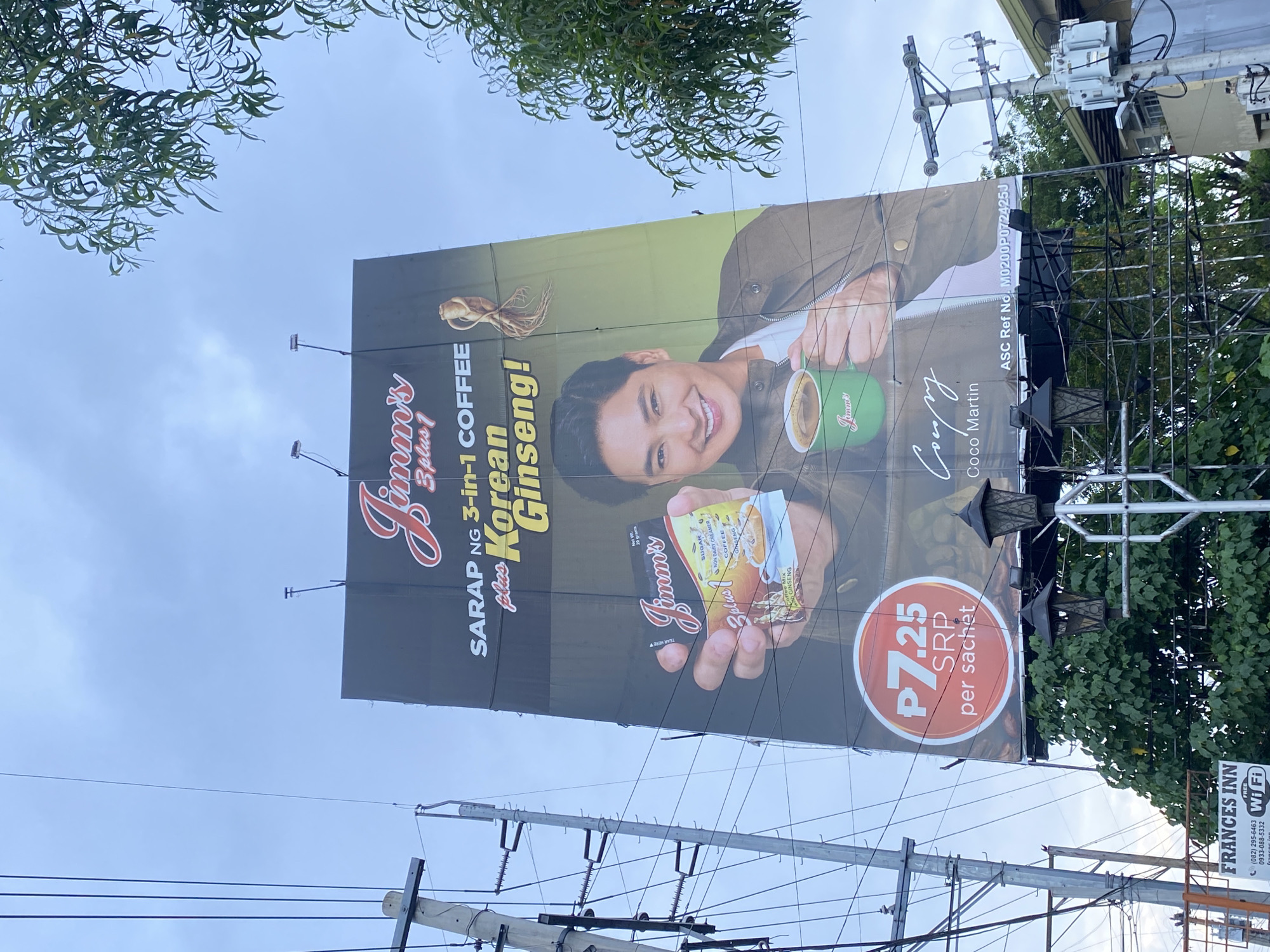 An outdoor billboard advertising "Jimmy's 3-plus-1 Coffee plus Korean Ginseng" is prominently displayed against a cloudy sky. The billboard features a close-up of a man, identified as Coco Martin, holding a green mug of coffee. He is smiling and wearing a brown jacket over a white t-shirt. In his other hand, he holds a small packet of Jimmy's coffee. The text on the billboard highlights "SARAP NG 3-in-1 COFFEE plus Korean Ginseng!" and a price of "P7.25 SRP per sachet." A root of Korean ginseng is depicted near the top right of the billboard. Below the main image, the signature "Coco Martin" is visible, along with an ASC Ref No. The billboard is supported by a metal structure and surrounded by trees and power lines. A street lamp is attached to the billboard structure. In the lower left corner of the image, a sign for "FRANCES INN FREE WIFI" is partially visible. The image is taken from a low angle, looking up at the billboard. The presence of the "Frances Inn" and the general urban setting suggests a location within Davao City, Philippines.