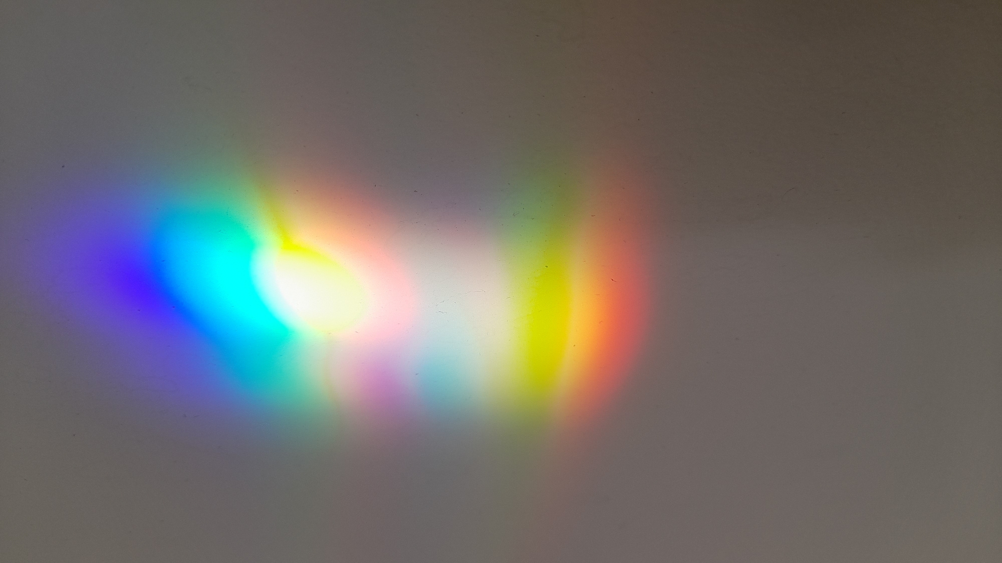 A blurry spectrum of light is projected onto a grey wall. The colors visible are purple, blue, cyan, green, yellow, orange, and red, proceeding sequentially across the wall. The intensity of the light fades as it spreads out from the center. There is no evidence to suggest any location, especially Saint Petersburg, Russia. No people, subjects, objects, actions, or interactions are apparent in the image, aside from the light and the wall it projects onto.