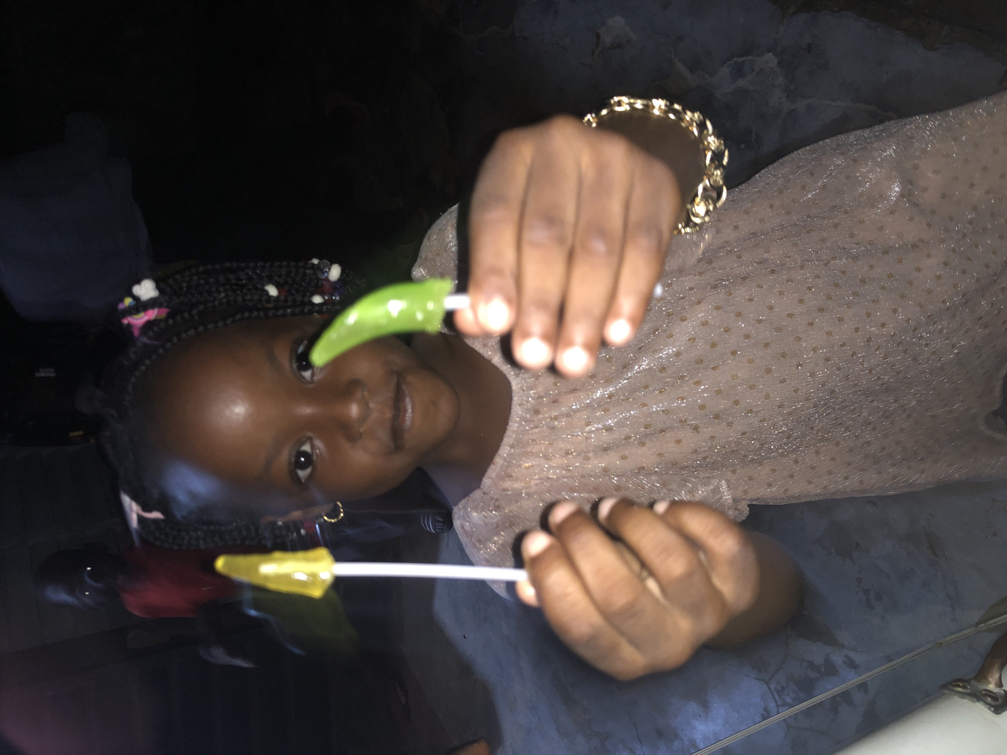 A young Black girl, with braided hair adorned with pink and white beads, stands indoors, holding two lollipops. One is yellow and the other is green. She is wearing a sparkly, light brown dress and a gold chain bracelet. In the background, another person, likely a child, is visible wearing a red shirt. The floor appears to be concrete. The lighting suggests it is nighttime. The image was taken in Ado, Nigeria.