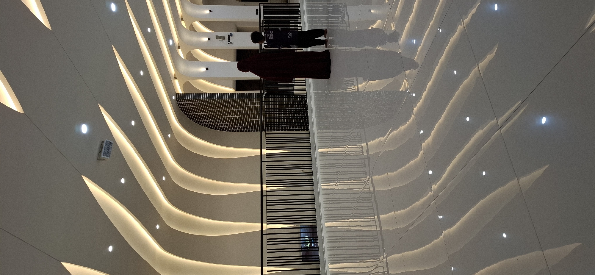 The image depicts an interior scene of a modern building, likely a public space or a place of worship given the architectural style and the attire of the individuals. In the foreground, a highly reflective floor shows distorted patterns of light originating from the ceiling. The ceiling itself features a striking design with curved, illuminated elements that create a wave-like effect.
Two individuals are visible in the mid-ground. On the left, a person dressed in a dark maroon abaya (a loose, outer garment) stands facing away from the camera. To their right, a child, also facing away, is dressed in a dark t-shirt with some text on the back and dark pants. Both individuals appear to be standing on an elevated walkway or balcony, separated from the foreground by a black metal railing.
Behind the railing, the architecture continues with vertical decorative elements and what appears to be a wall clad in dark, gridded tiles. The overall lighting is dim, with the primary illumination coming from the concealed lights within the ceiling's curves, casting a warm, ambient glow. The environment suggests a contemporary and possibly elegant setting. The location is identified as Karangpandan, Indonesia.