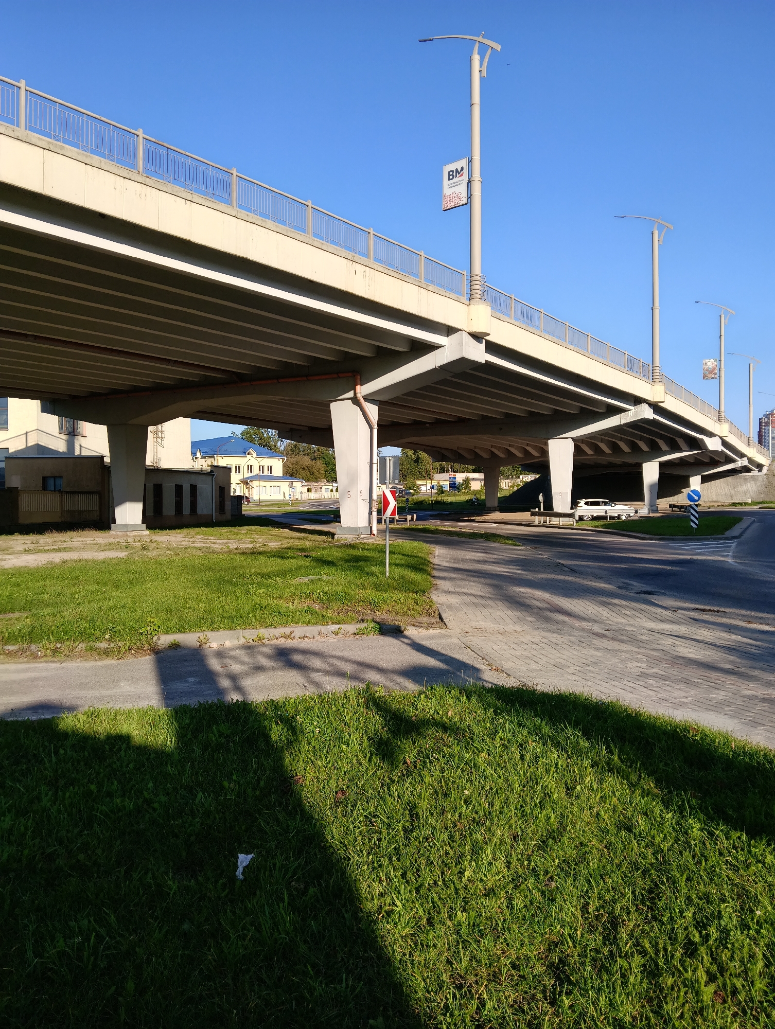 The image shows an underpass of an overpass in Hrodna, Belarus. The overpass is made of concrete and has a metal railing on top. There are several concrete pillars supporting the overpass. On the left side of the image, there are some buildings visible, including a yellow building with a blue roof. On the right side of the image, there is a road with a white car parked under the overpass. There are also streetlights and traffic signs visible. The ground is covered with grass and pavement. The sky is clear and blue.
