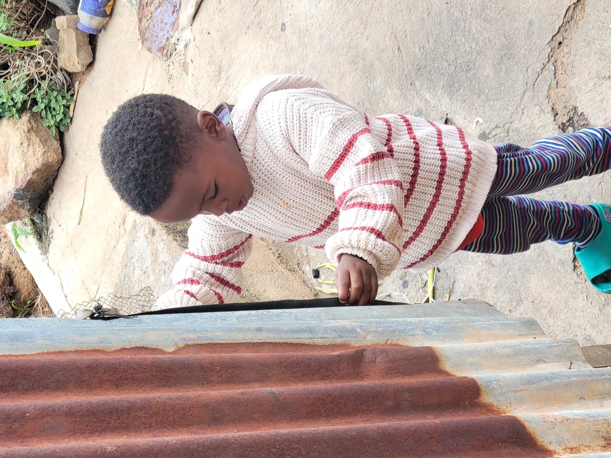 A young child lies prone on a corrugated metal sheet. The sheet is rusted on one side and grey on the other. The child has dark, closely cropped hair and wears a beige and maroon striped sweater, striped pants and a green sandal. The child's hand rests on a black rubber strip on the corrugated sheet. Behind the sheet is a concrete or stone surface with some vegetation. The location is Jos, Nigeria.