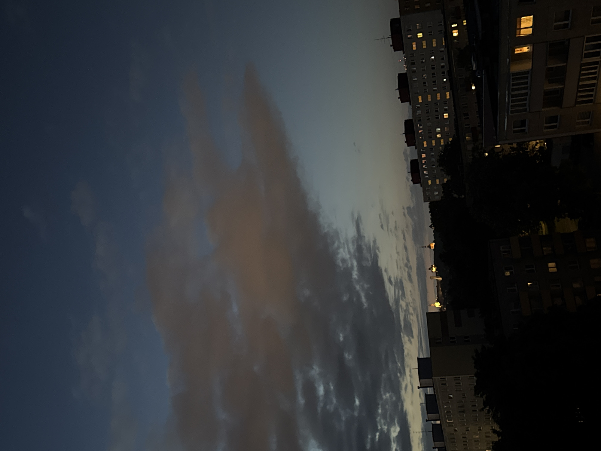 An elevated nocturnal view of an urban landscape in Tallinn, Estonia, features residential buildings in the foreground and midground, with illuminated historical architecture on a distant hill. The sky, characteristic of late dusk or night, transitions from dark blue-grey in the upper left to a deeper blue-black in the upper right, partially covered by extensive, dense grey and reddish-brown clouds.
The lower two-thirds of the image are dominated by multiple multi-story residential buildings, primarily in dark grey and beige tones, with numerous windows emitting warm yellow light. These structures exhibit flat rooftops, some with visible utility units or antennae. Dark, silhouetted tree masses are interspersed among the buildings, particularly in the lower-left and central portions of the frame. No individuals, vehicles, or discernible activities are visible in the foreground or midground.
In the distant background, positioned on an elevated landform (Toompea Hill), a cluster of at least three distinct historical architectural structures is prominently illuminated with artificial yellow-orange light. These features, including what appears to be an onion dome and spires, are identifiable elements of Tallinn's Old Town. The scene is static, devoid of any apparent movement or interaction.