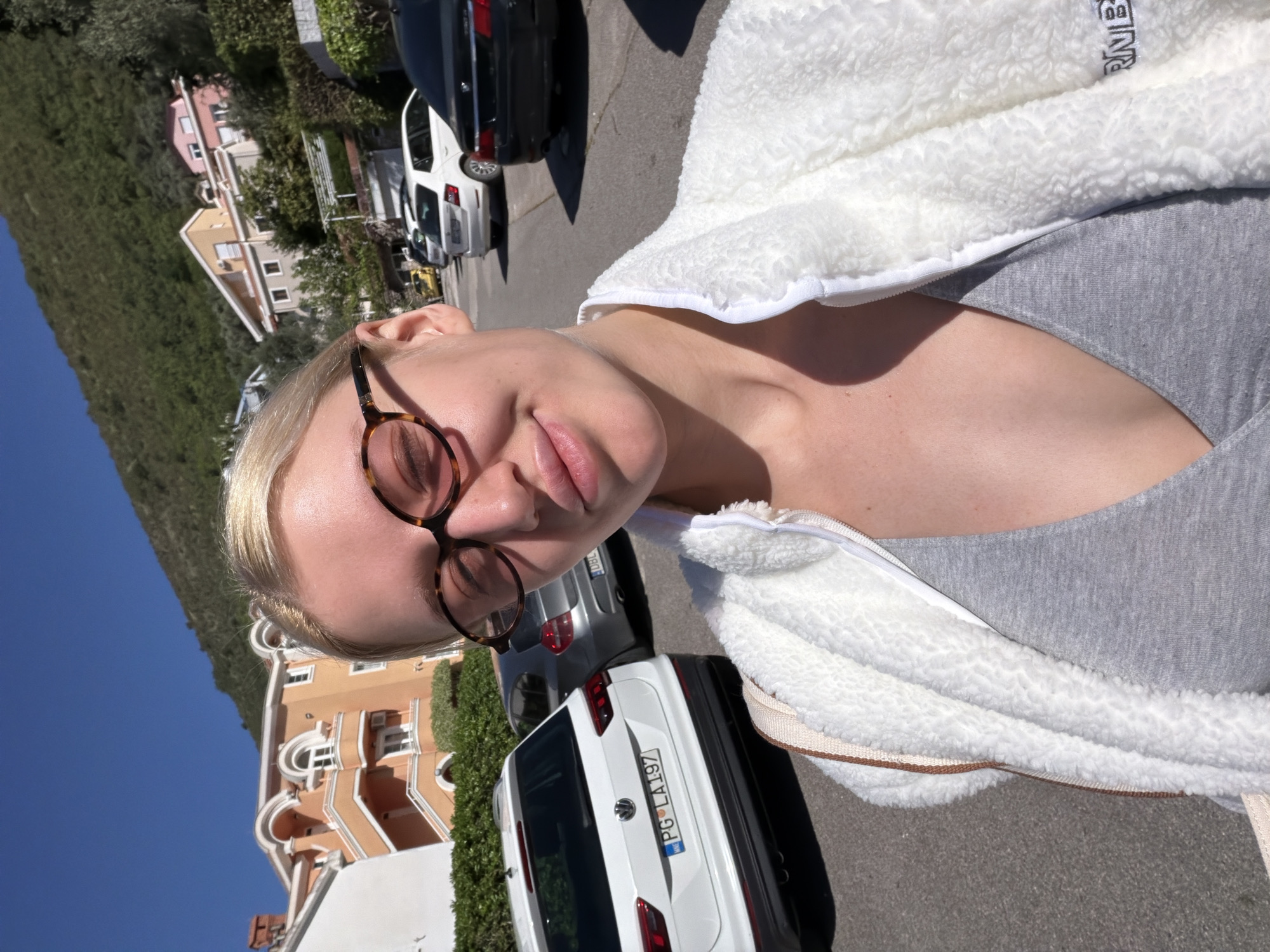 A woman is posing for a selfie outdoors on a sunny day in what appears to be Nerin, Montenegro. She is wearing a gray top, a white fleece jacket, and tortoiseshell glasses. The background features a hillside with green foliage, residential buildings with terracotta roofs, and several parked cars. A white car with the license plate "PG LA197" is in the foreground.