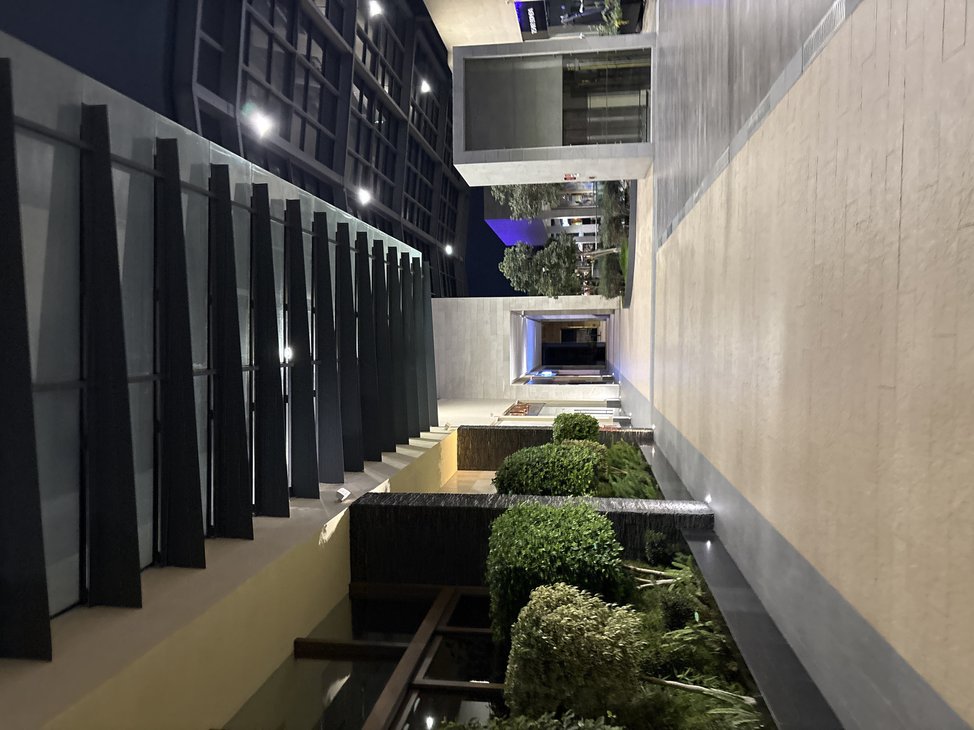 The photograph depicts a modern architectural space, likely an outdoor corridor or public area, in Puebla, Mexico. The perspective is a low-angle, looking upwards and along the length of the space.
On the left, a tall building facade with vertical black slats fronts the area. A glass barrier runs along the building. The right side features a light-colored concrete wall, with a gray band at the top.
Below these structures, a series of landscaped terraces ascend toward the center of the image. These terraces are filled with green bushes and trees. A thin stream of water is falling down from one terrace to the next.
In the background, a paved area extends, with other modern buildings visible. A blue colored structure can be seen among the trees. Overhead lighting is present throughout the space, and it appears to be nighttime.
The composition and architectural style suggest a contemporary urban environment.