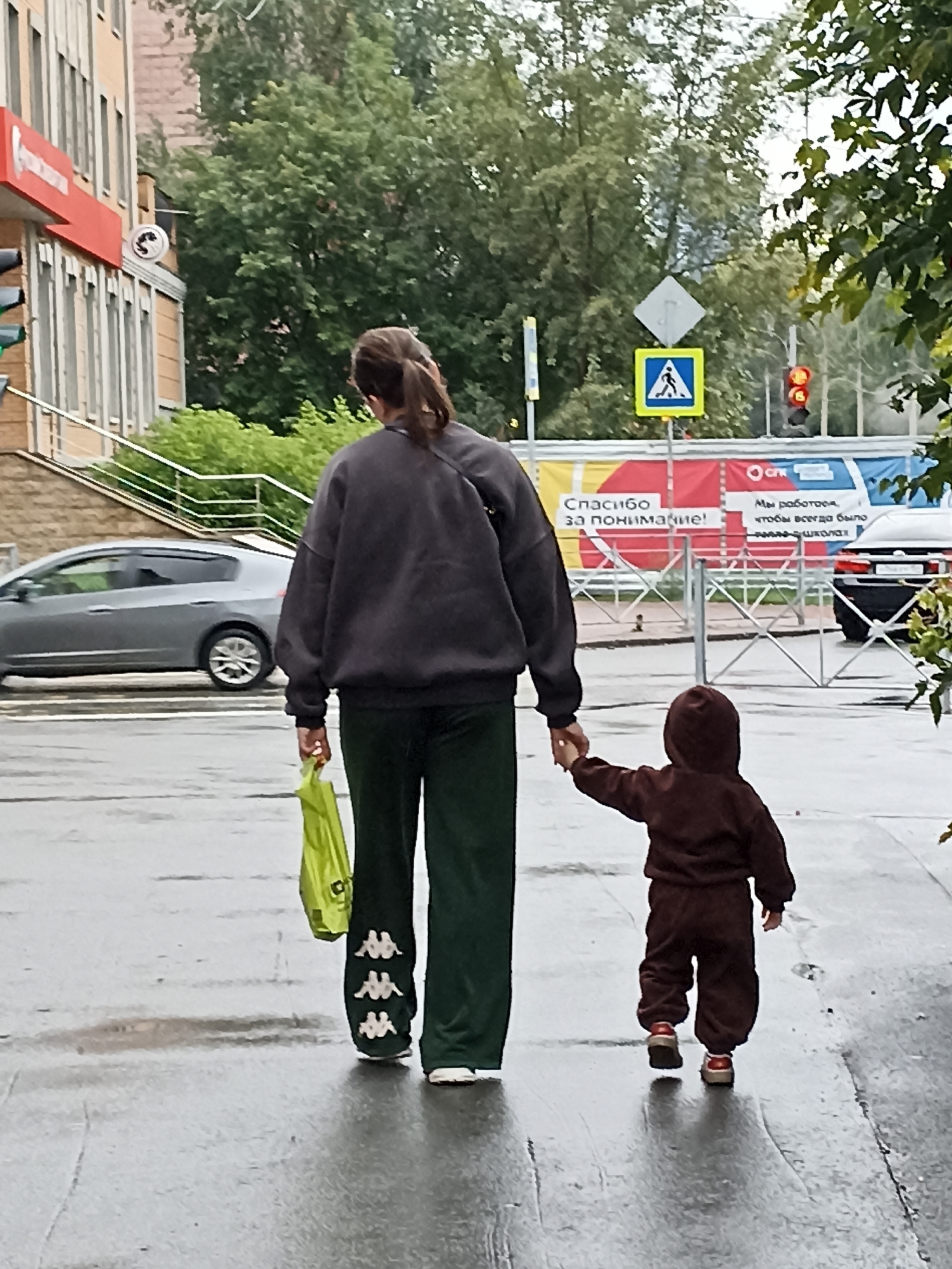 The image shows two figures walking away from the camera on a wet paved surface, likely a sidewalk. The figure on the left is an adult, appearing to be a woman, with dark hair pulled back in a ponytail. She is wearing a dark grey oversized sweatshirt and dark green sweatpants with white Kappa logos. She is holding a bright yellow plastic bag in her left hand. The figure on the right is a small child, wearing a brown hooded tracksuit and red-accented shoes. The adult and child are holding hands.
In the background, a grey sedan is parked on the left. Behind the figures, there is a metal fence and a large banner. The banner has text that appears to be in Russian, with sections in yellow, red, and blue. The text on the banner includes "СПАСИБО ЗА ПОНИМАНИЕ!" (Thank you for your understanding!) and "МЫ РАБОТАЕМ, ЧТОБЫ ВСЕГДА БЫЛО - ШКОЛА!" (We are working so that there is always - school!).
Further in the background, there are trees and a building with red and beige elements. There are also traffic lights and street signs, including a pedestrian crossing sign. The overall environment suggests an urban setting. The wet pavement indicates recent rainfall or damp conditions. While the image does not explicitly state the location, the Russian text on the banner strongly suggests the scene is in Russia. Given the request to include the city of Novosibirsk, Russia, it is plausible that this image was taken there.