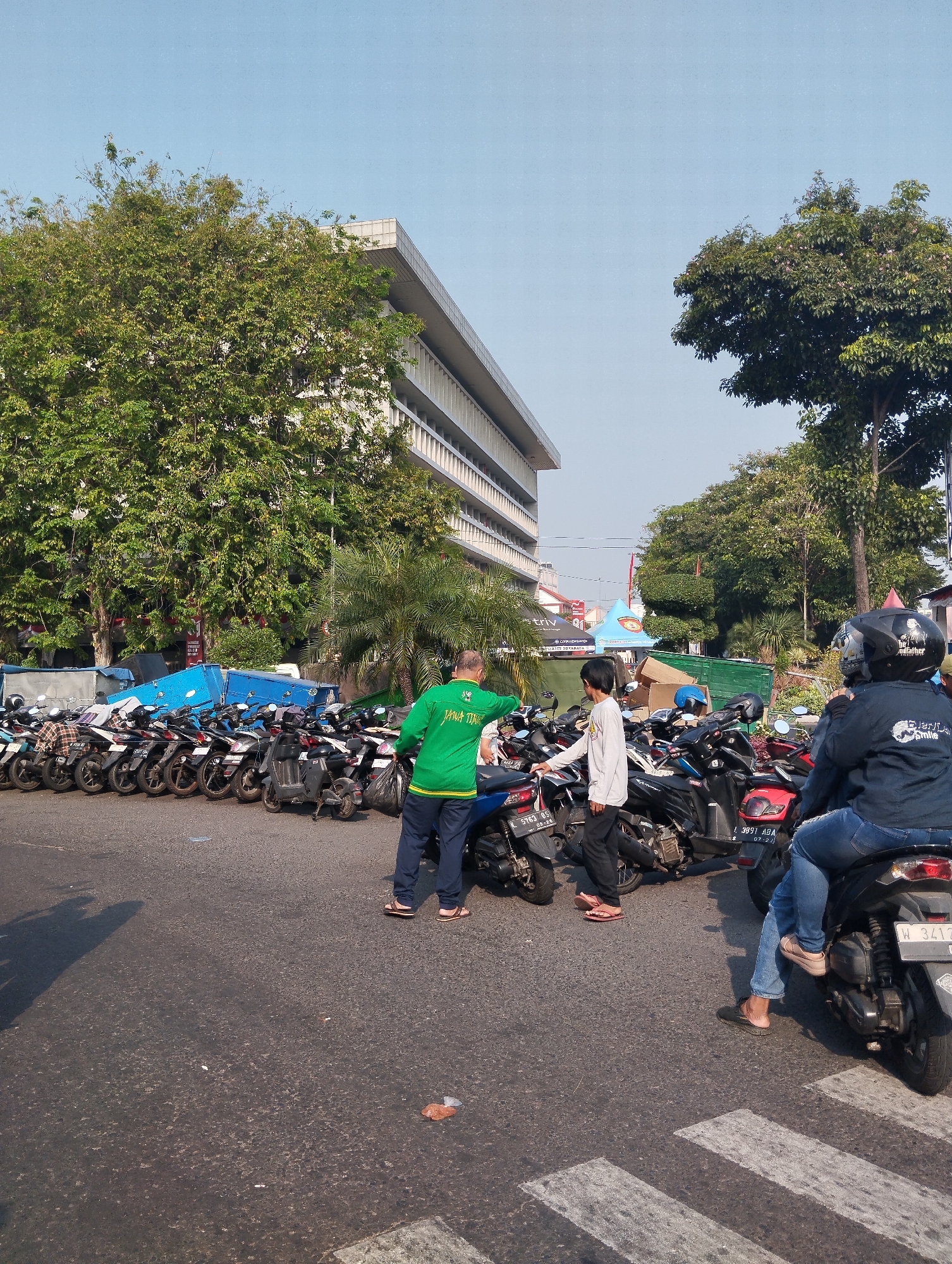 The image depicts a street scene in Surabaya, Indonesia, likely a parking area outside a large building. In the foreground, a dark asphalt road with white pedestrian crossing lines is visible. Several motorbikes are parked in rows, occupying a significant portion of the frame.
Three individuals are interacting in the middle ground. One person, wearing a bright green long-sleeved shirt with yellow trim and dark pants, is holding a black trash bag. Another person, wearing a white long-sleeved shirt and dark pants, is standing near the motorbikes. A third person, wearing a dark jacket and jeans, is seated on a motorbike in the right foreground, wearing a helmet. The motorbikes are of various models and colors, with some displaying license plates.
In the background, a modern, multi-story building with white columns and a light-colored facade dominates the scene. Lush green trees, including a large one in the left middle ground and a palm tree, are interspersed with the building. Several blue portable structures and a green truck are also visible behind the motorbikes. The sky is clear and blue, indicating a sunny day. The overall impression is one of a busy urban environment, possibly a public space or the entrance to a facility.